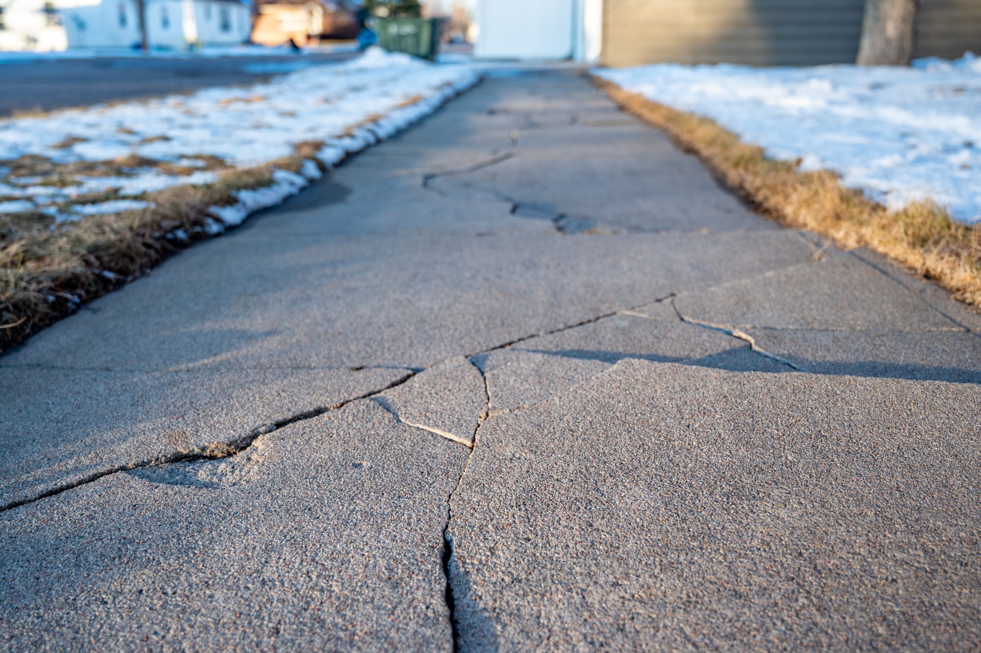 A close-up low shot of a very cracked sidewalk flanked by snow-covered brown grass.