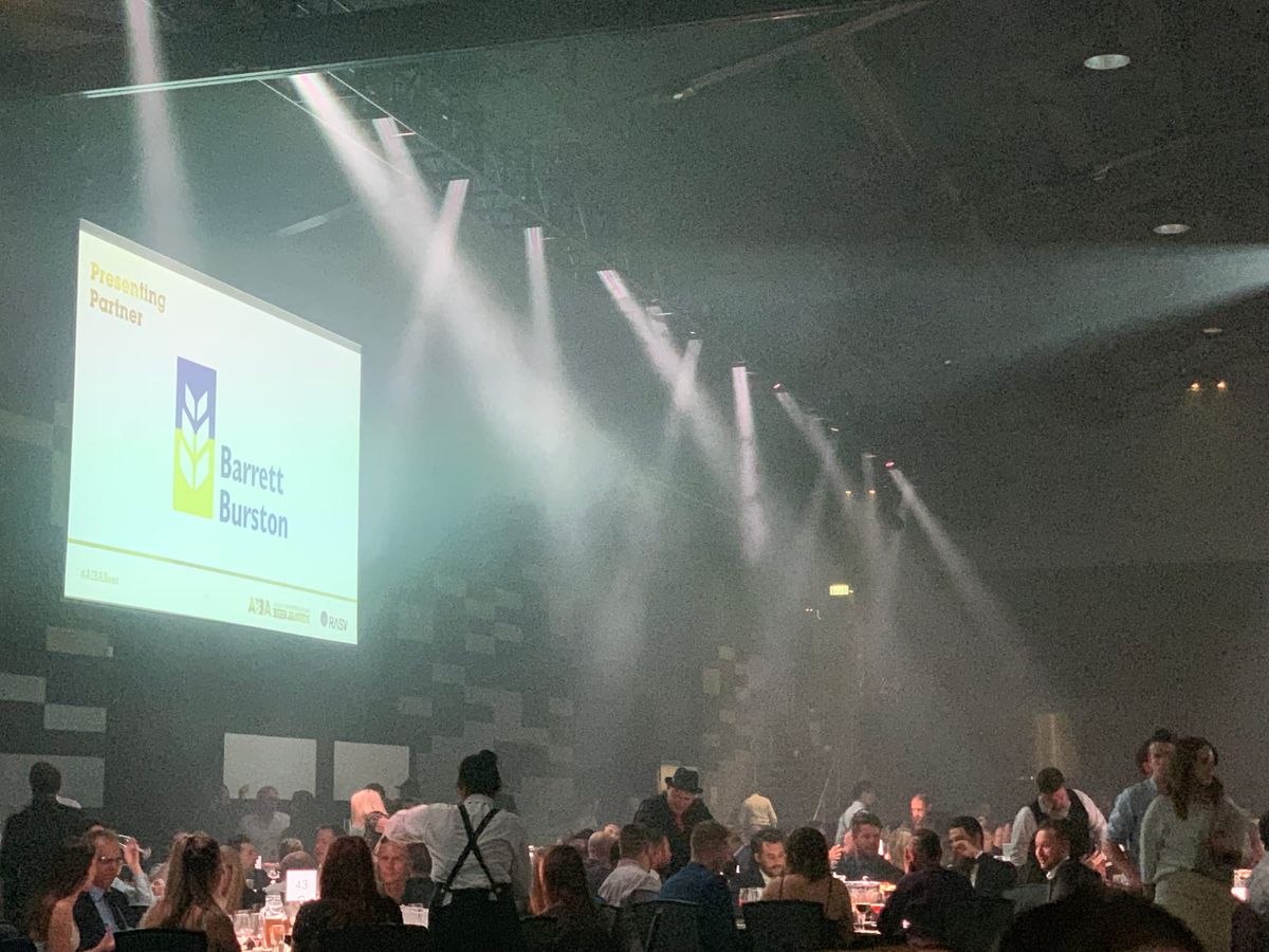 A large screen at an awards dinner showing the Barrett Burston logo above the words "Presenting Partner", with stage lighting and a crowded room of seated guests below.