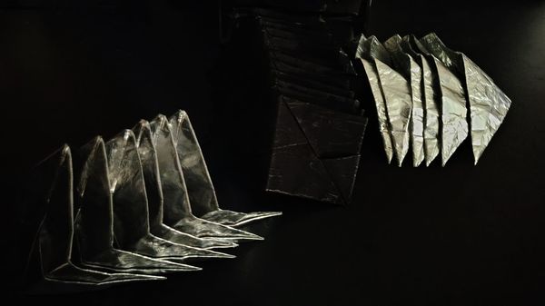 Twelve matte black parallelograms folded such that they have three segments rest in a pile flanked on either side by a pile of six reflective parallelograms similarly folded.