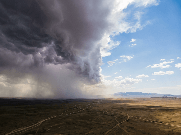 Dark thunderstorm clouds with lightning meet clear blue sky above dry open land with winding dirt roads