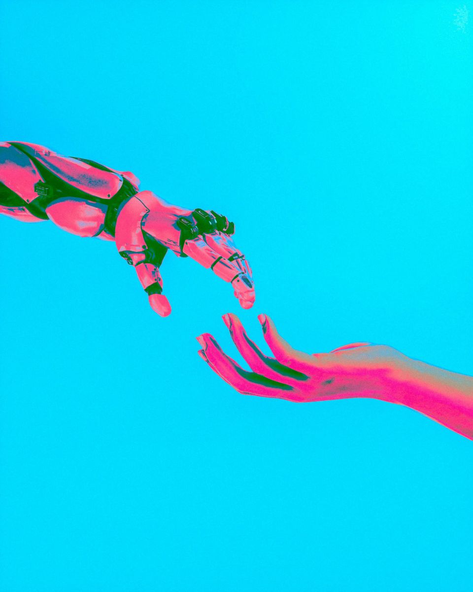 Two pink hands; one belonging to a human, one to a robot, are almost touching, against a turquoise background.   