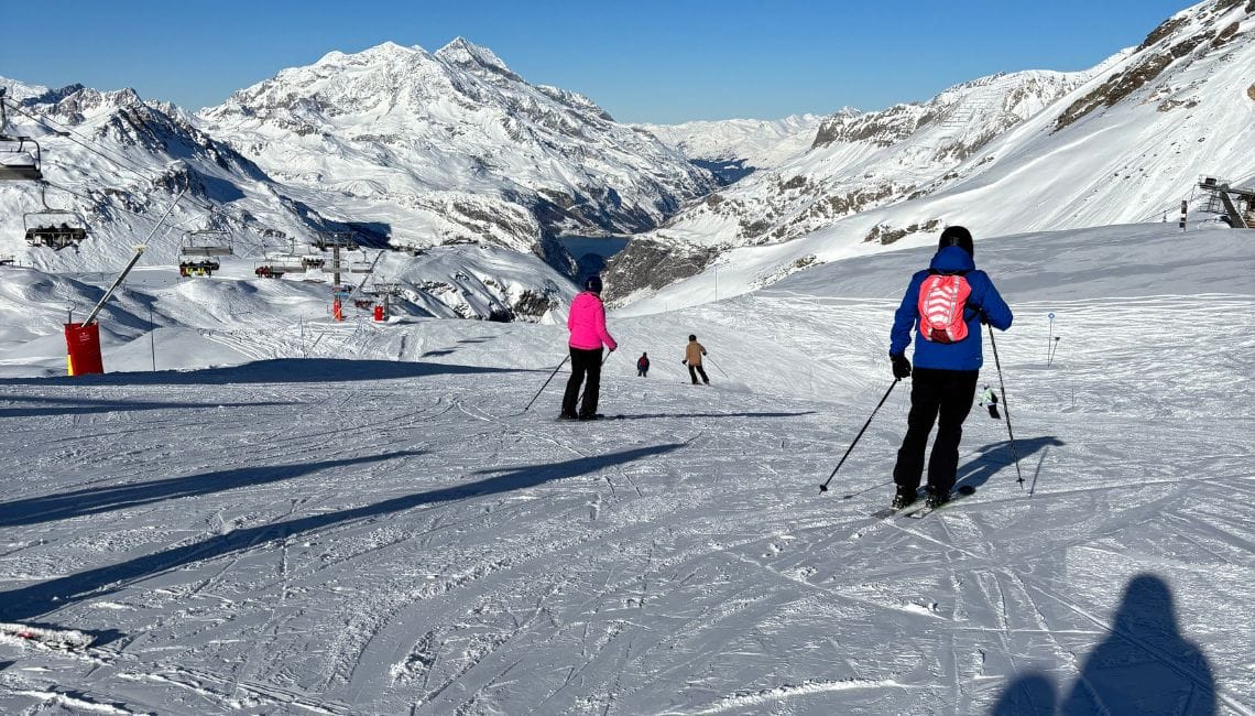 Skiers going down a shallow gradient slope with dramatic mountain background and chairlift to left