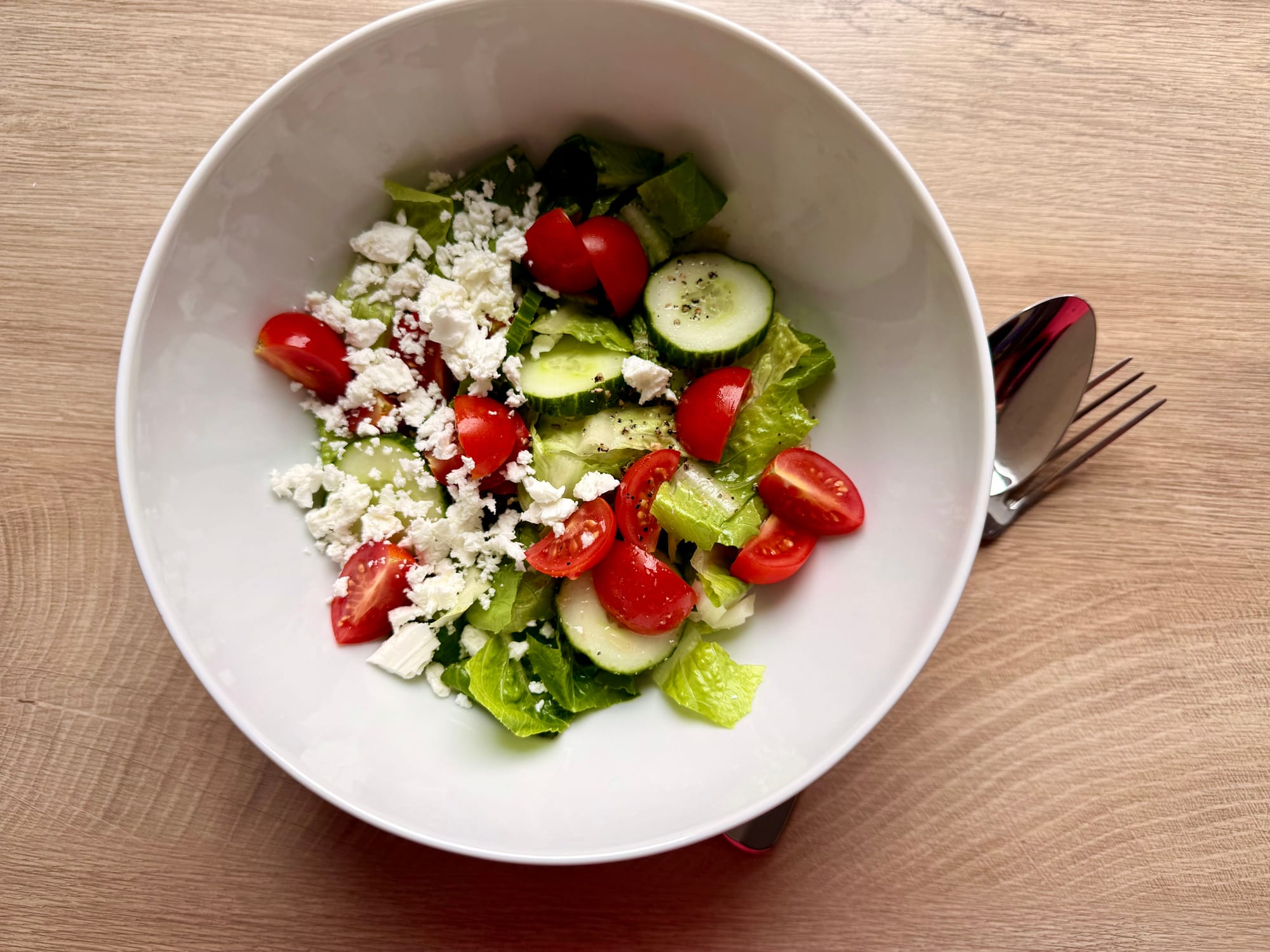 Draufsicht auf eine weiße Schüssel mit einem frischen Salat aus geschnittenem Römersalat, Gurkenscheiben, halbierten Kirschtomaten und weißem Feta-Käse, daneben Silberbesteck.
