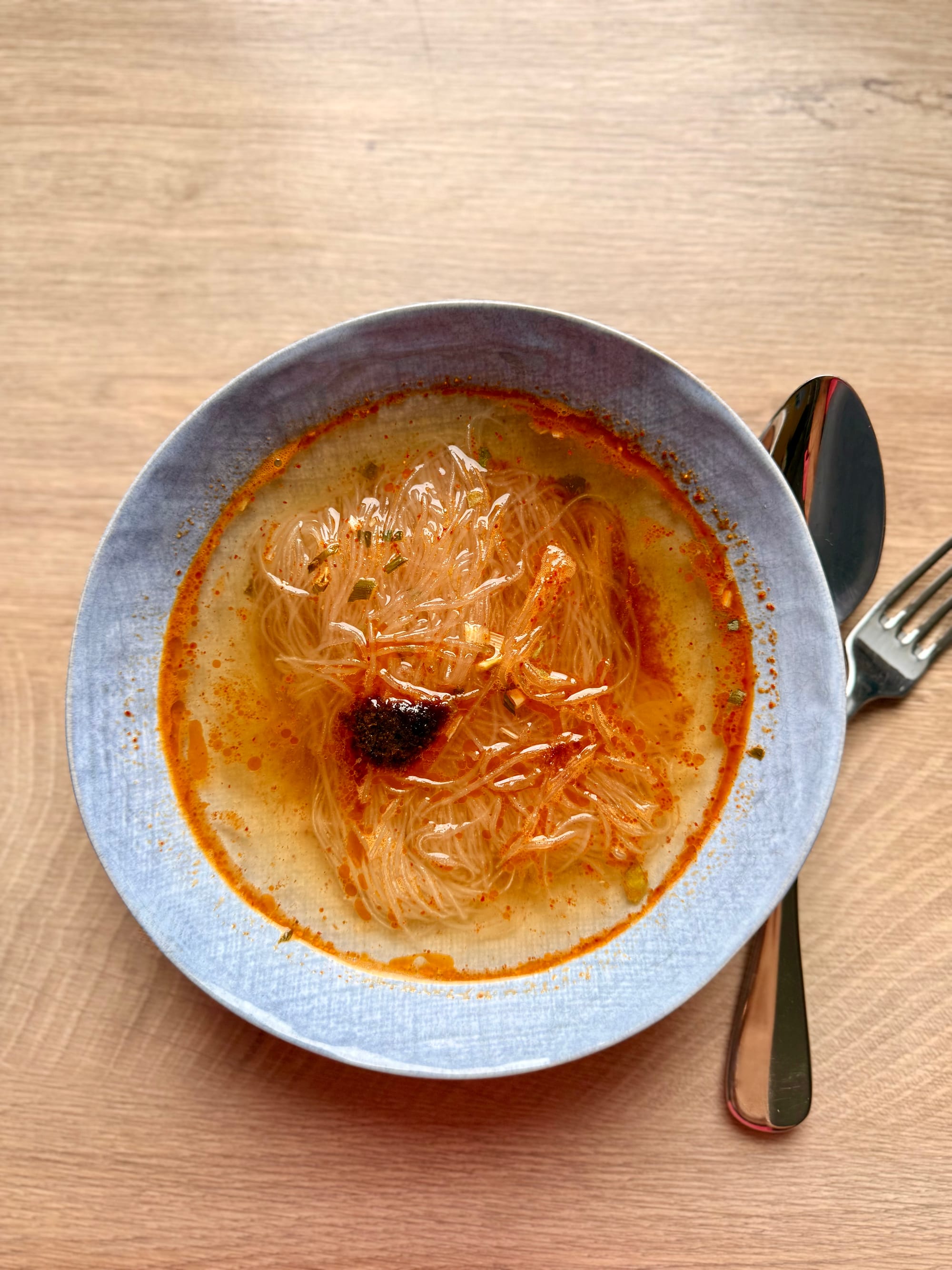 Draufsicht auf eine blaue Keramikschüssel mit Instant-Glasnudelsuppe in einer rötlich-orangen Brühe, serviert mit Löffel und Gabel.