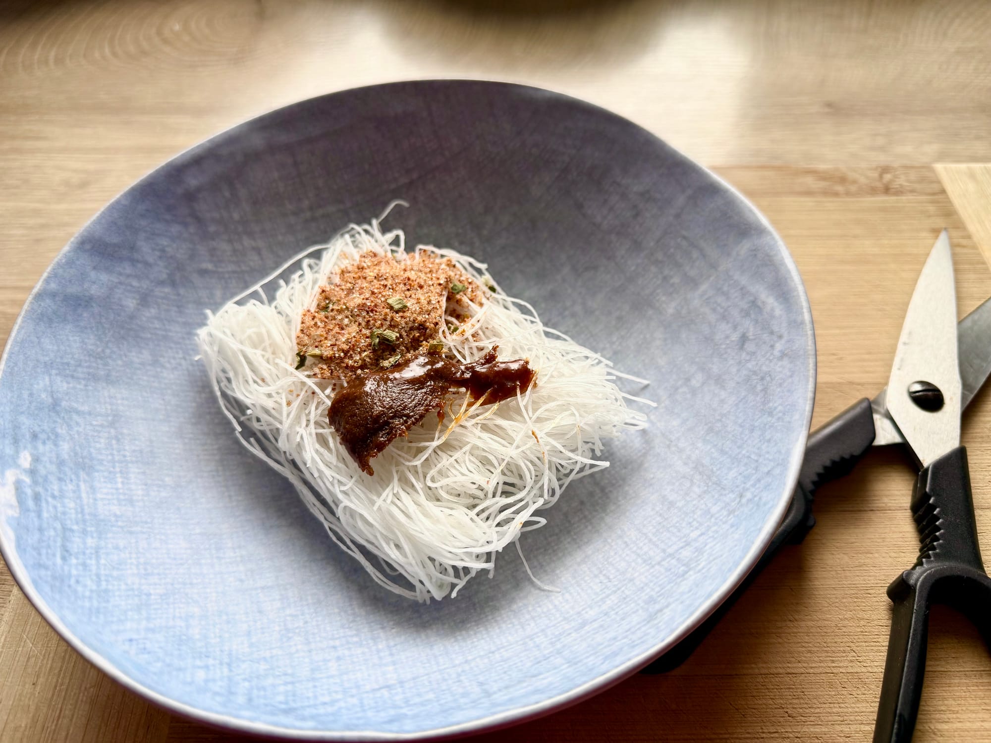 Close-up of dry instant rice vermicelli noodles topped with reddish spice powder and dark seasoning paste inside a light blue bowl.