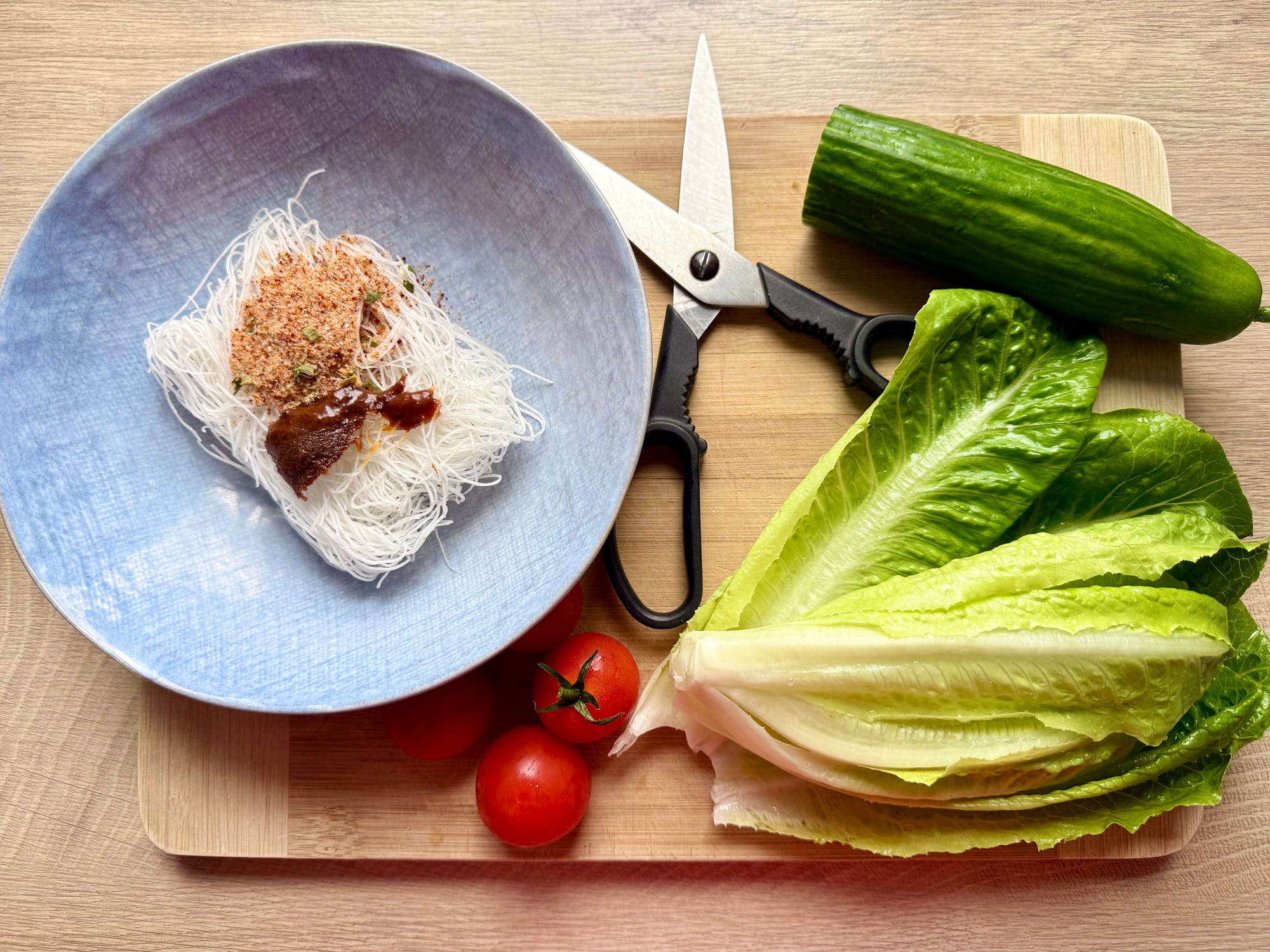 Nahaufnahme eines Holzschneidebretts mit einer Schüssel trockener Instant-Nudeln und Würzpaste links, einer Küchenschere in der Mitte sowie frischem Römersalat und einer Gurke rechts.
