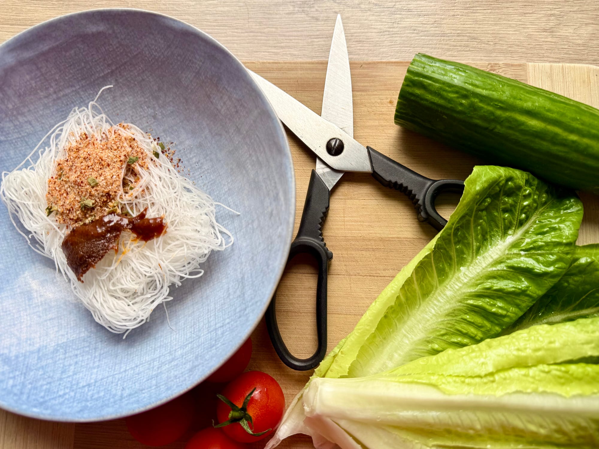 A close-up view of a wooden cutting board with a bowl of dry instant noodles and flavor paste on the left, kitchen scissors in the center, and fresh romaine lettuce with a cucumber on the right.