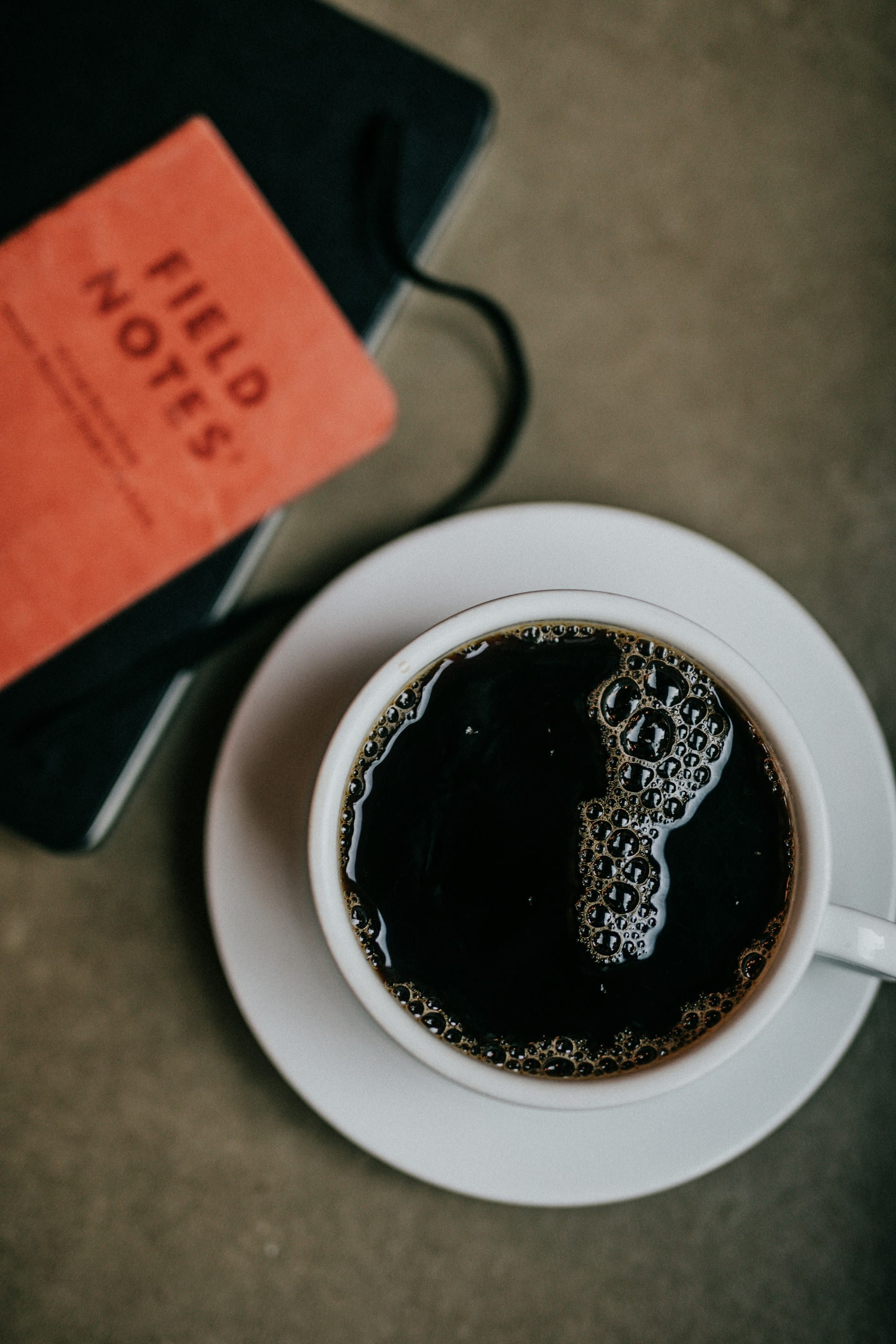 EN: Top-down view of a white cup filled with black coffee next to an orange Field Notes pocket memo book resting on a dark desk.