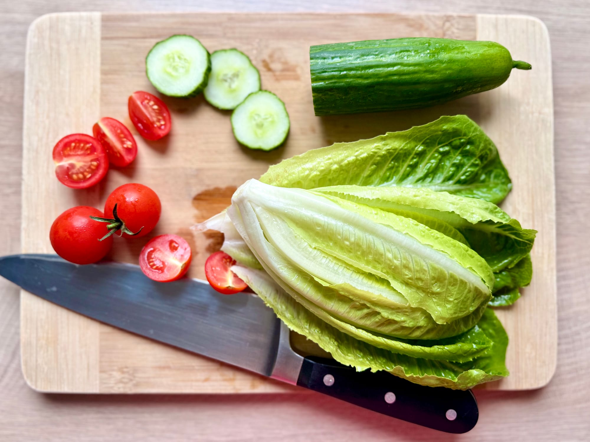 Draufsicht auf geschnittene Gurken, halbierte Kirschtomaten und frischen Römersalat, angerichtet auf einem Holzschneidebrett neben einem Küchenmesser.