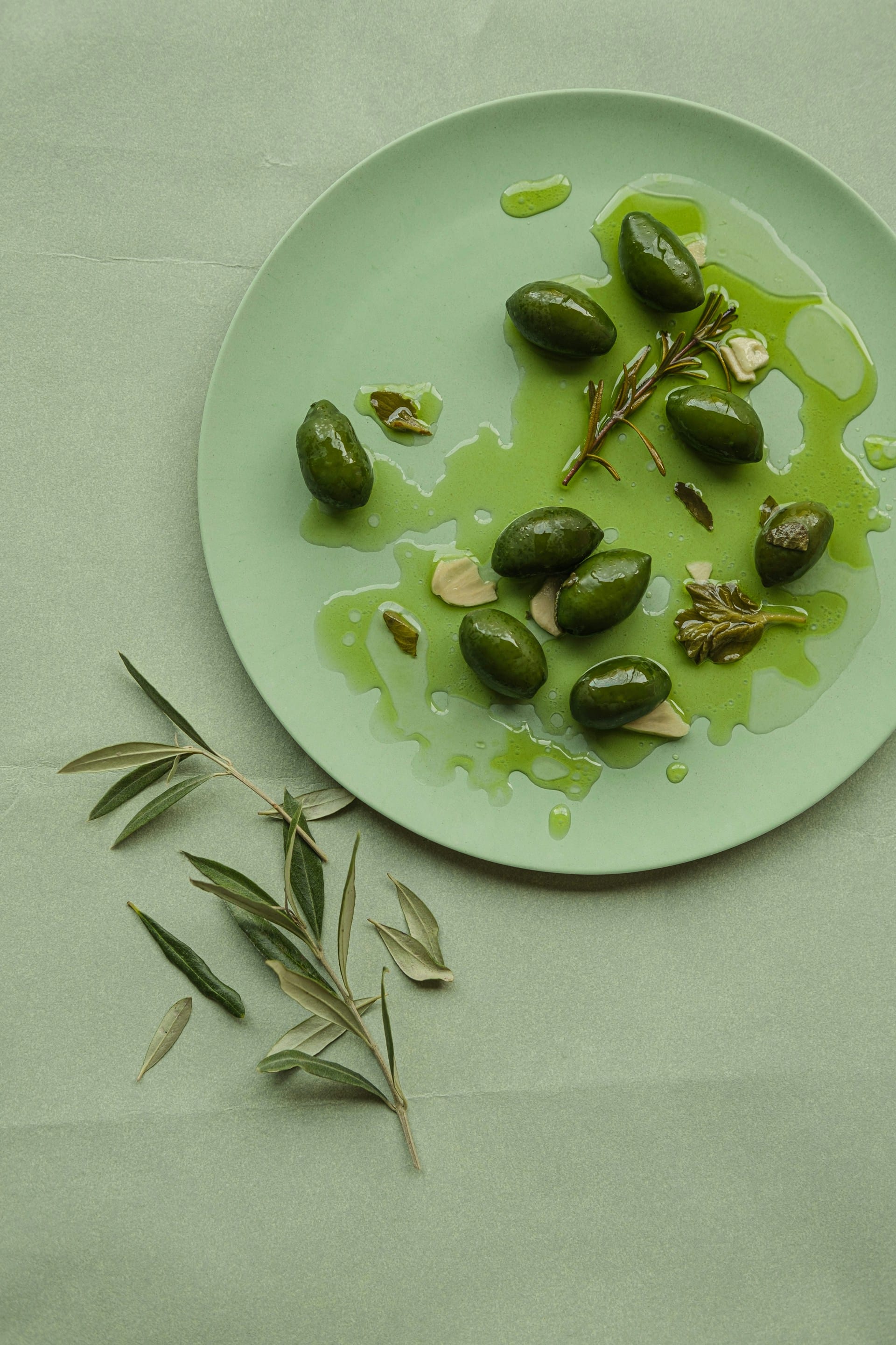 Top-down view of bright green olives drizzled with vibrant olive oil, garlic, and fresh rosemary sprigs on a sage green plate, accompanied by an olive branch.