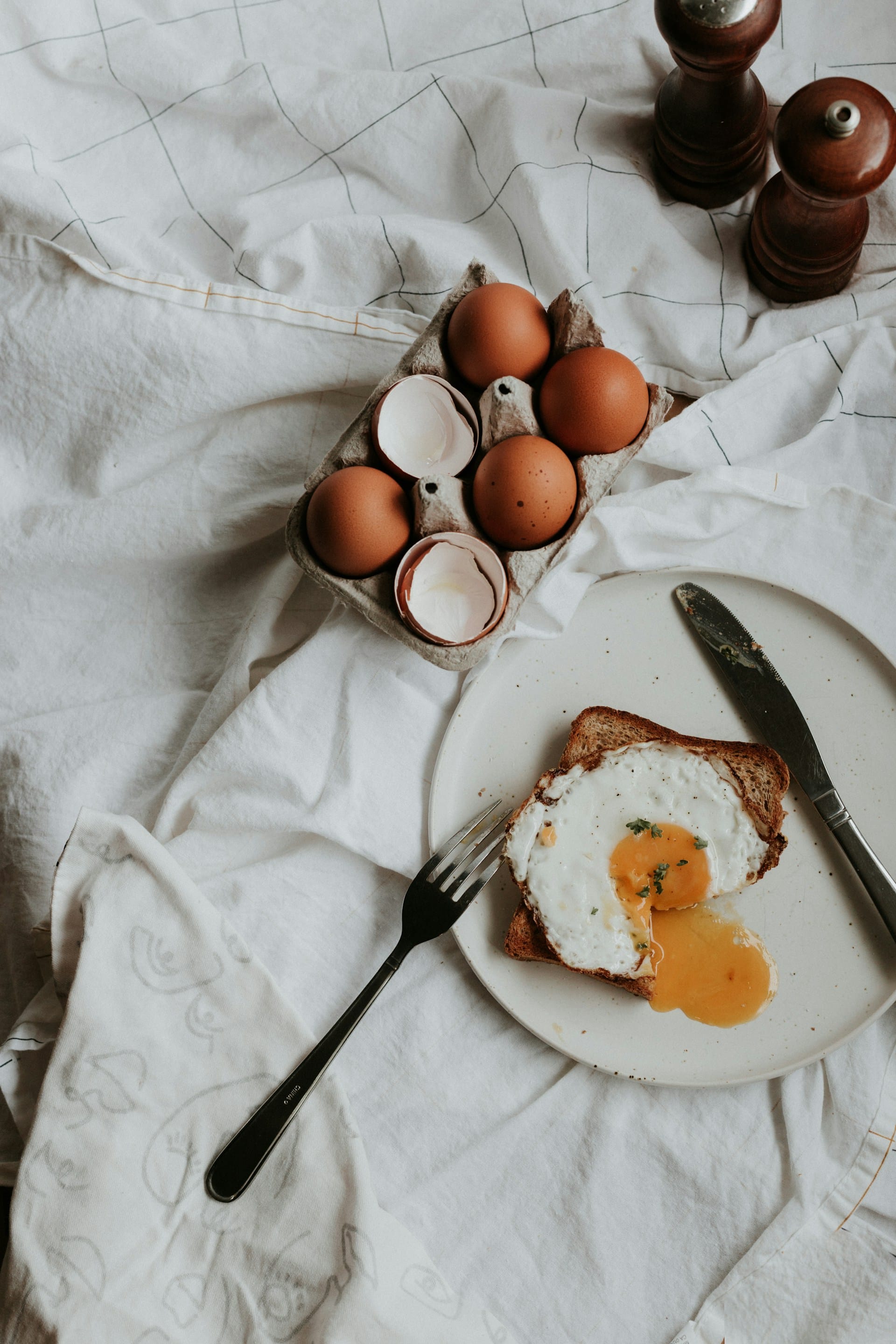 Flat-lay image of a sunny-side-up fried egg on a slice of toasted bread with a runny yellow yolk spilling onto a ceramic plate, surrounded by an egg carton with brown eggshells and wooden salt and pepper shakers.