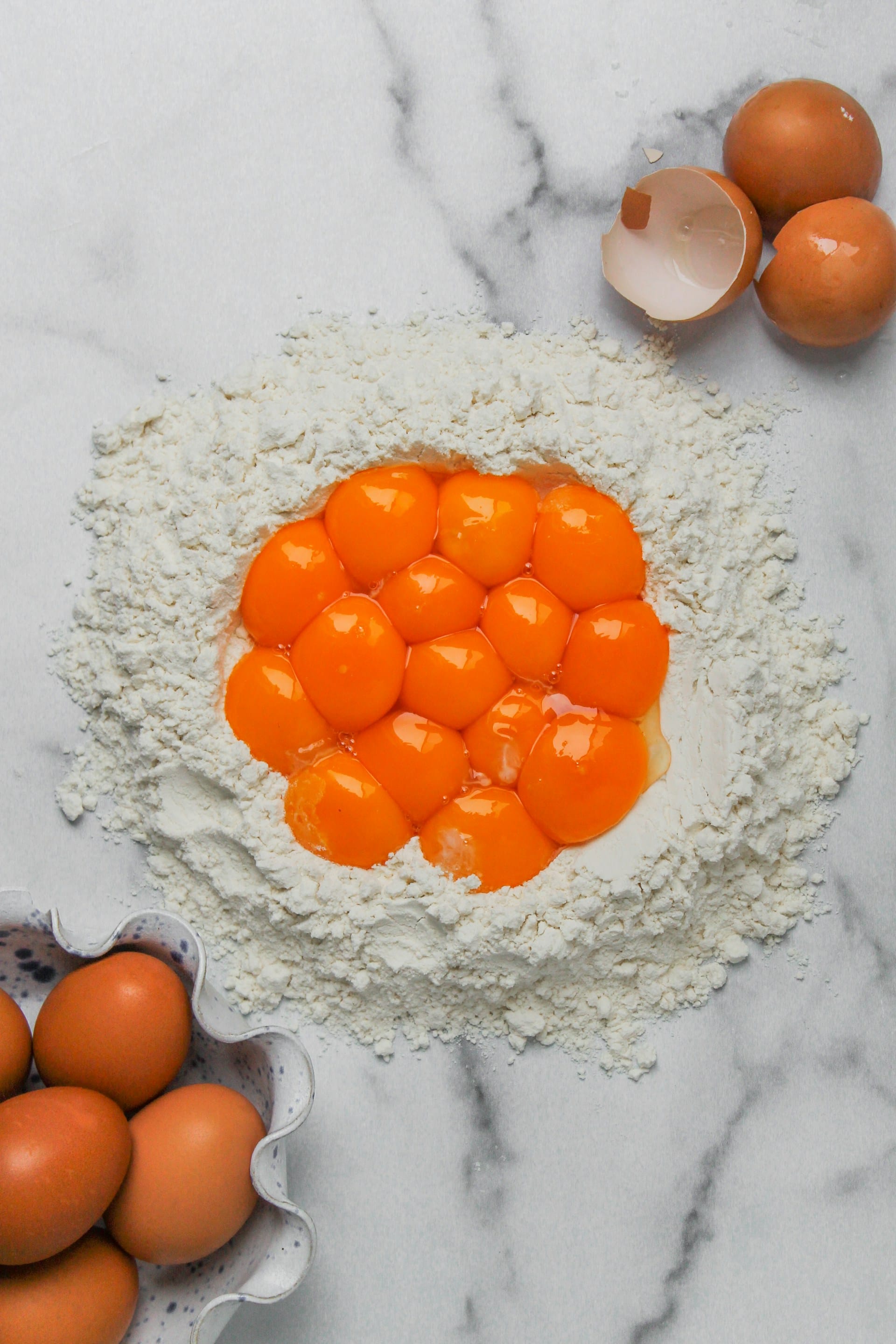 Top-down view of a nest of flour on a white marble countertop filled with twelve vibrant orange raw egg yolks, flanked by whole brown eggs and cracked shells.