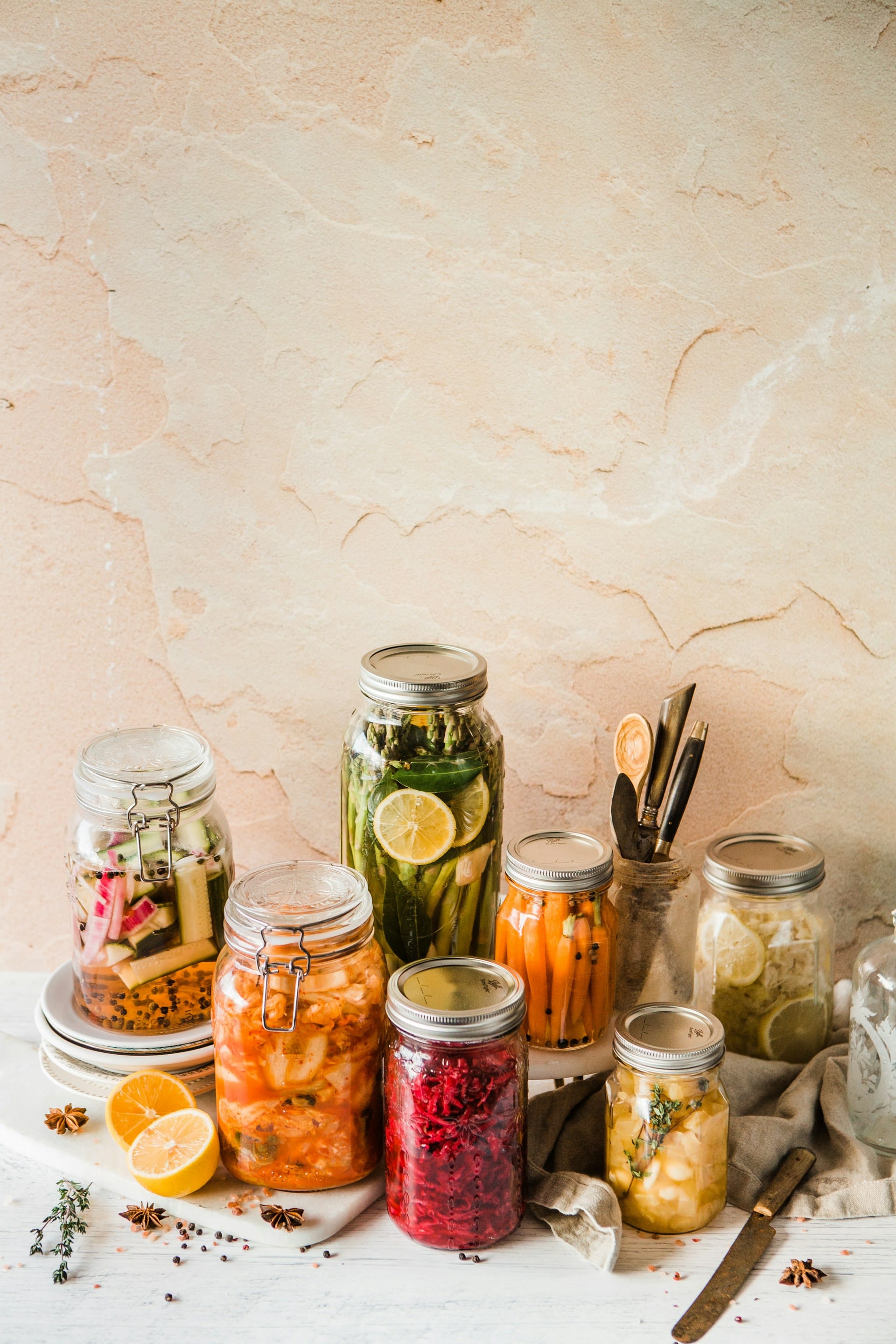 Assortment of glass mason jars filled with brightly colored fermented vegetables, including red cabbage, carrots, asparagus, and citrus, arranged on a light stone surface.