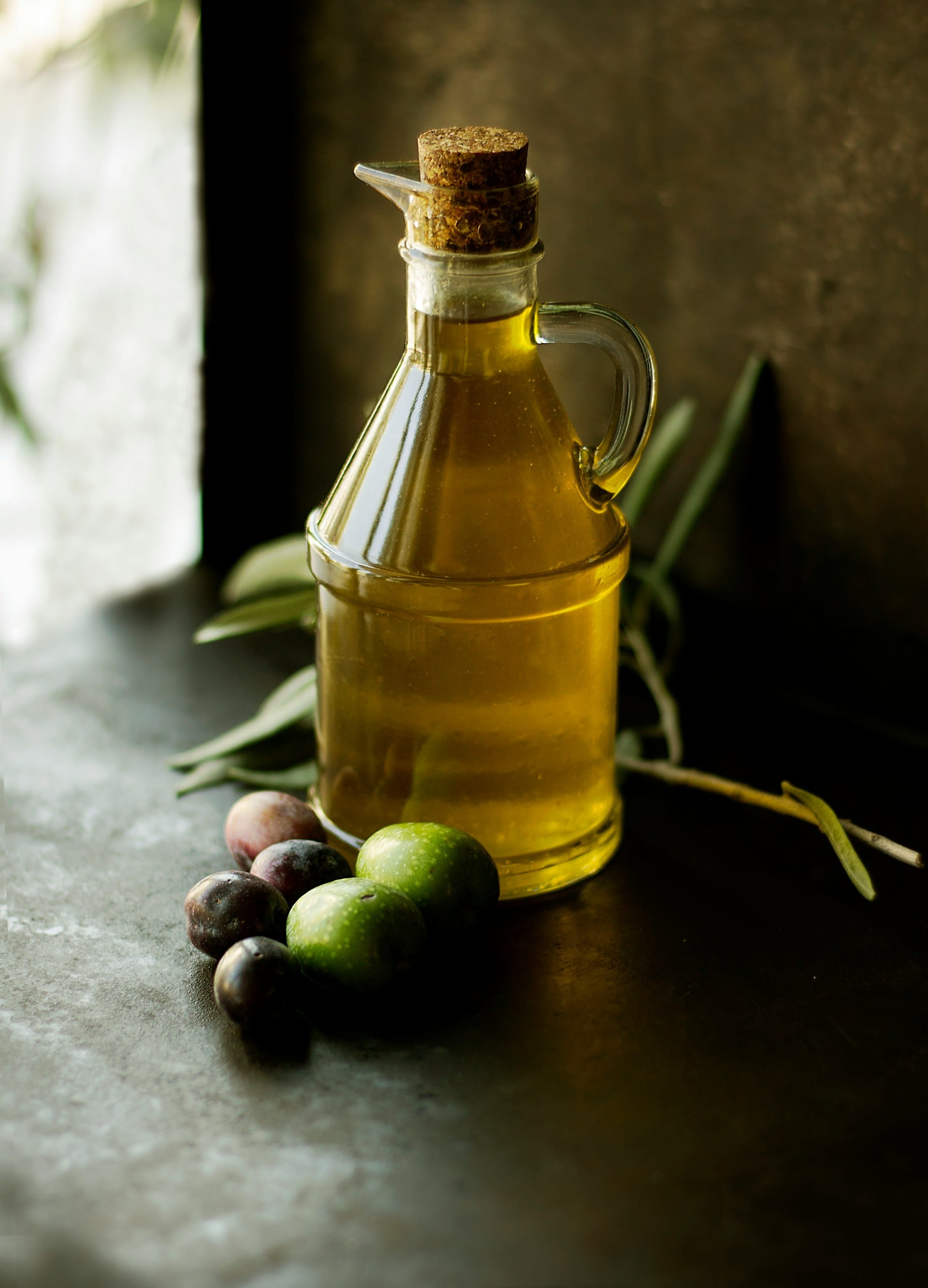 Eine kleine Glaskaraffe gefüllt mit goldenem Olivenöl, verschlossen mit einem Korken, stehend neben frischen ganzen Oliven und Olivenzweigen auf dunklem Untergrund.