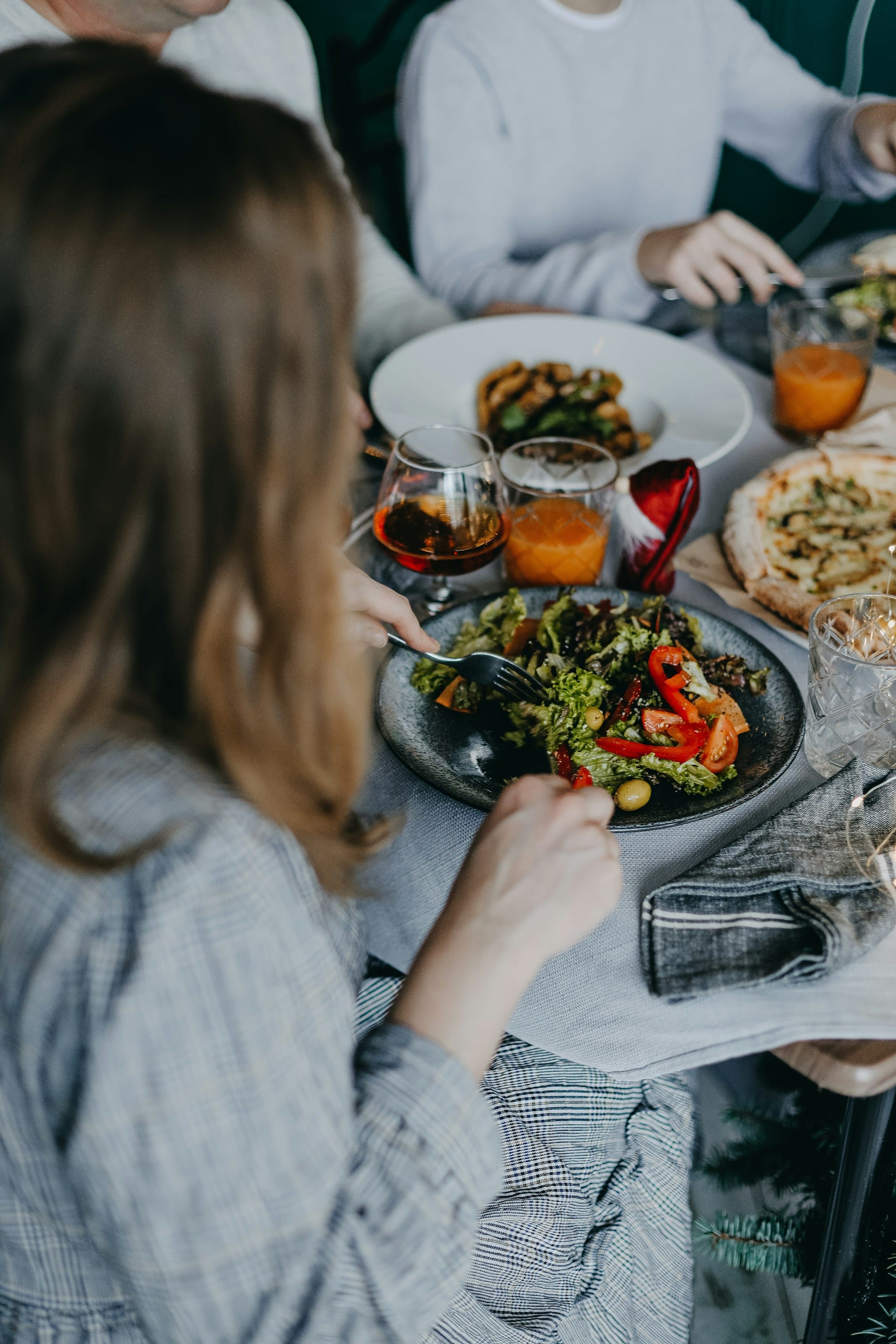 Over-the-shoulder view of family or friends seated around a dining table sharing fresh salads, juices, and warm dishes together.