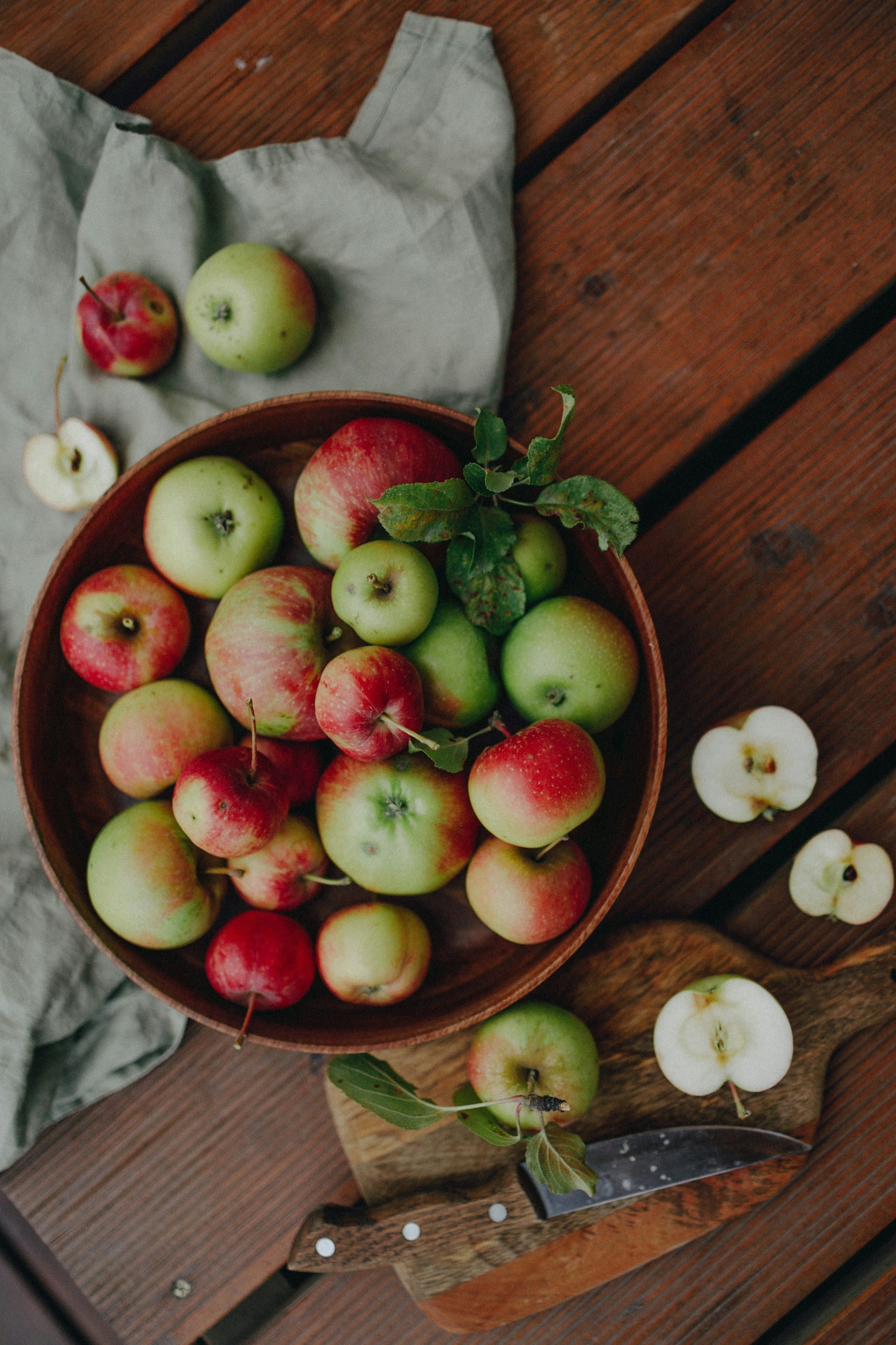 Top-down view of a round wooden bowl filled with various red and green garden apples next to sliced apple halves on a wooden surface.