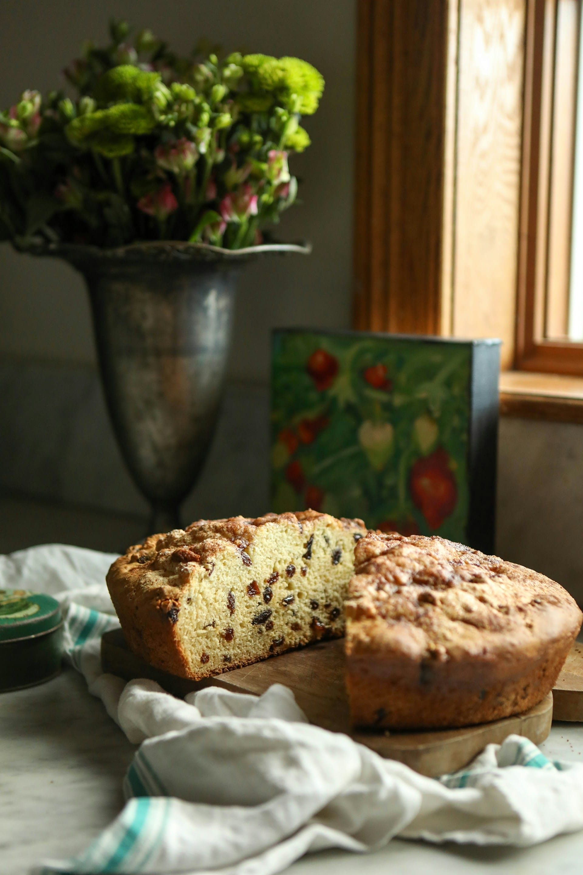 A round, golden-brown barmbrack fruit bread sliced open on a wooden board next to a vase of green flowers in a bright kitchen window.