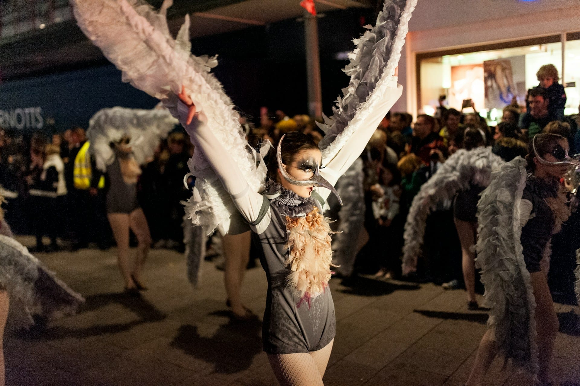A street performer wearing an ornate white feather bird mask and winged costume during a night parade surrounded by onlookers.