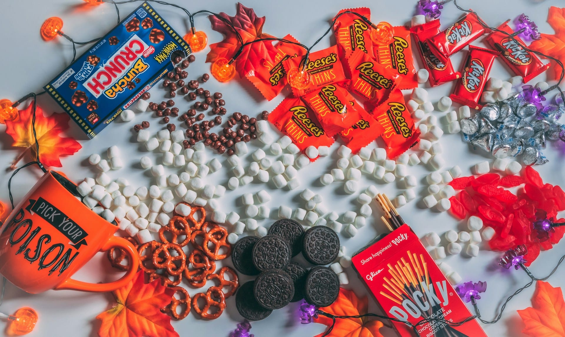 Flat-lay view of brightly wrapped commercial Halloween chocolates, candies, marshmallows, and pretzels spread out around a mug that reads "Pick Your Poison."