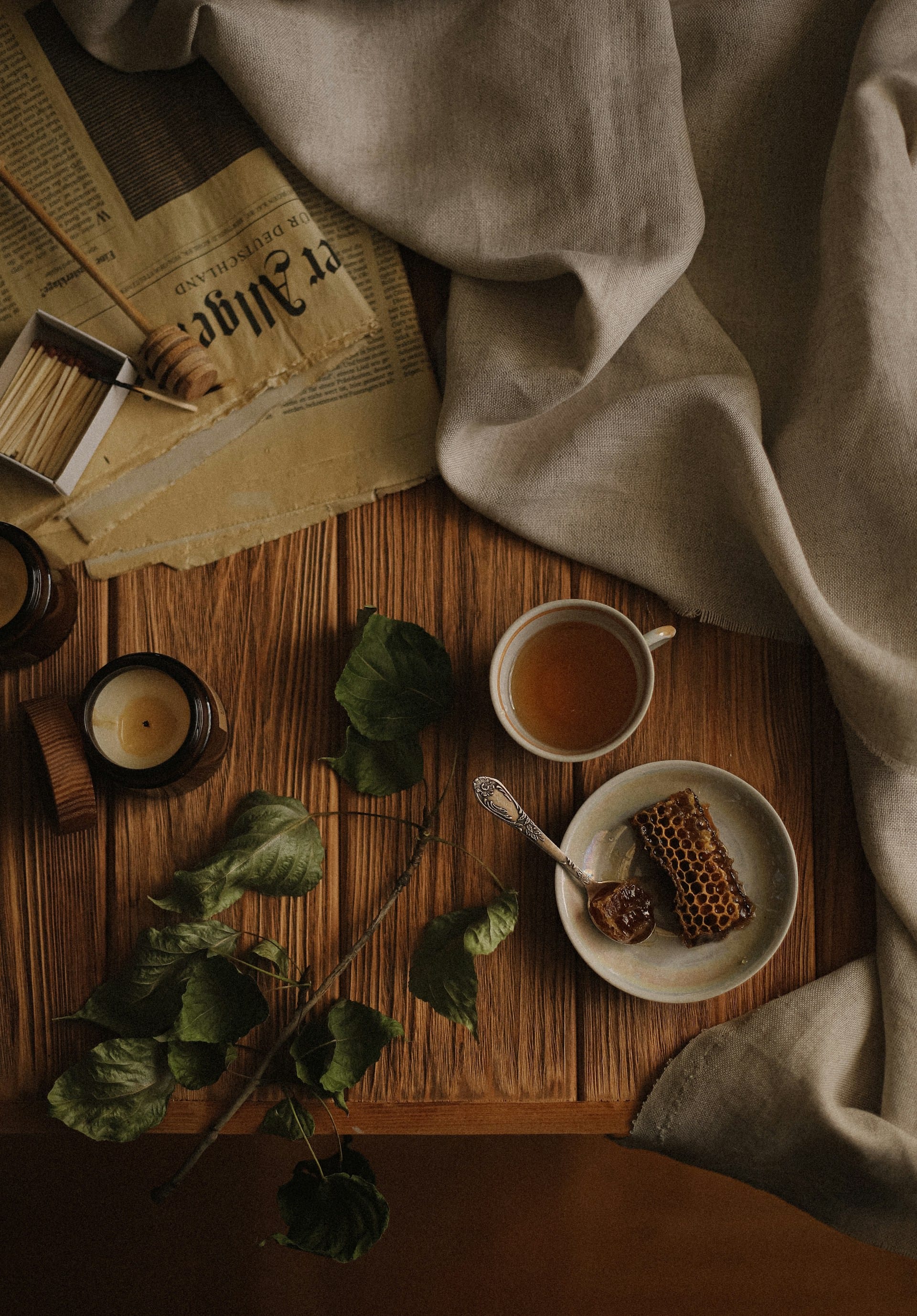 Moody overhead view of a rustic wooden table featuring a ceramic plate with a piece of raw honeycomb, a silver spoon, a cup of warm tea, green leaves, a lit candle, and vintage newspaper pages.