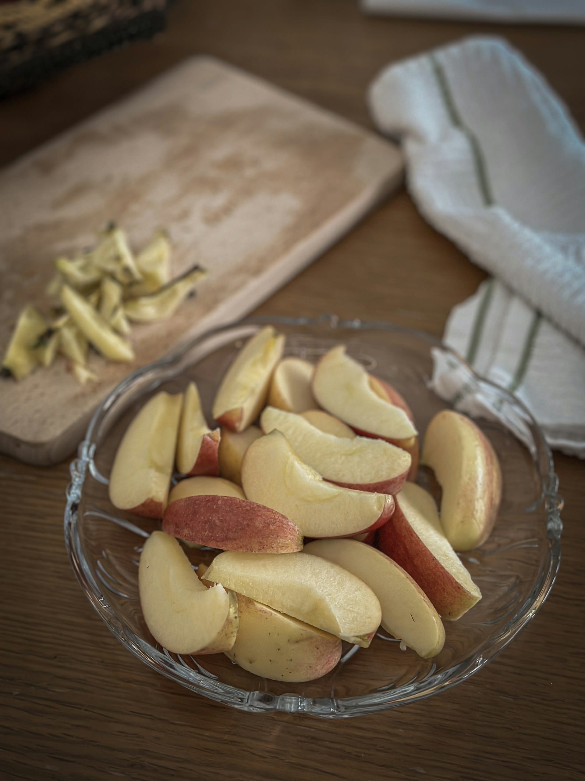 A glass dish filled with unpeeled, fresh apple wedges with a cutting board and apple cores in the soft-focused background.