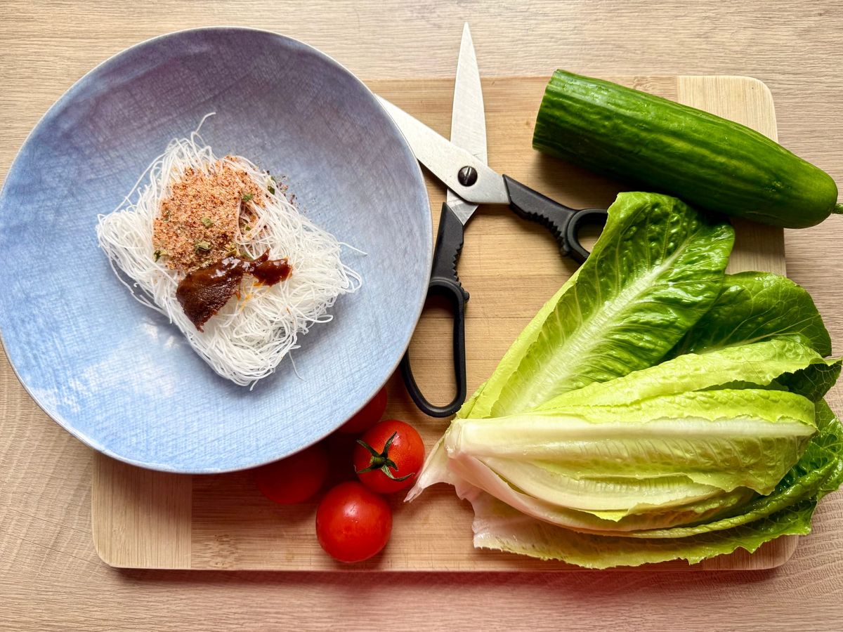 Flat-lay view of a wooden cutting board with dry instant glass noodles in a blue bowl alongside fresh romaine lettuce, cherry tomatoes, and a cucumber.