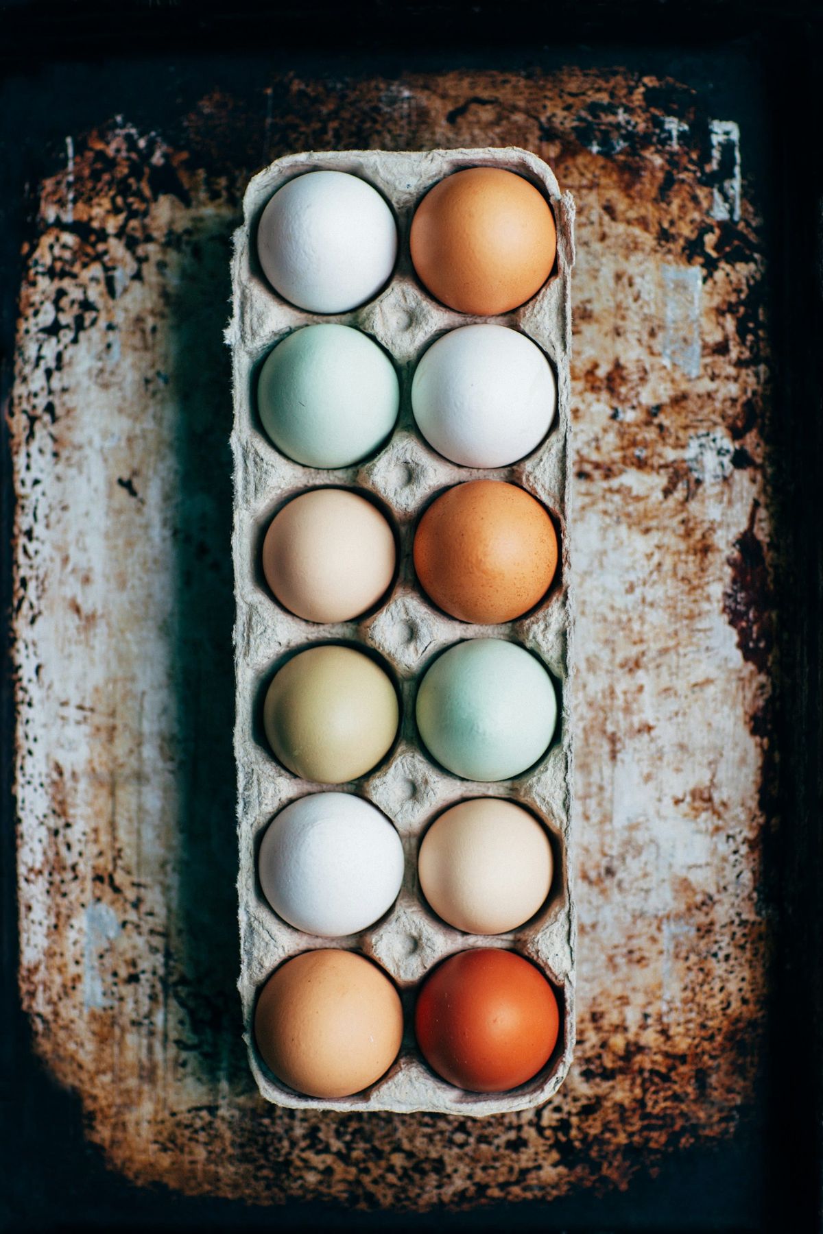 Top-down view of a dozen eggs in a cardboard carton resting on a rusty vintage baking sheet, displaying varying shell shades from white and cream to soft blue, pale green, and deep terra-cott