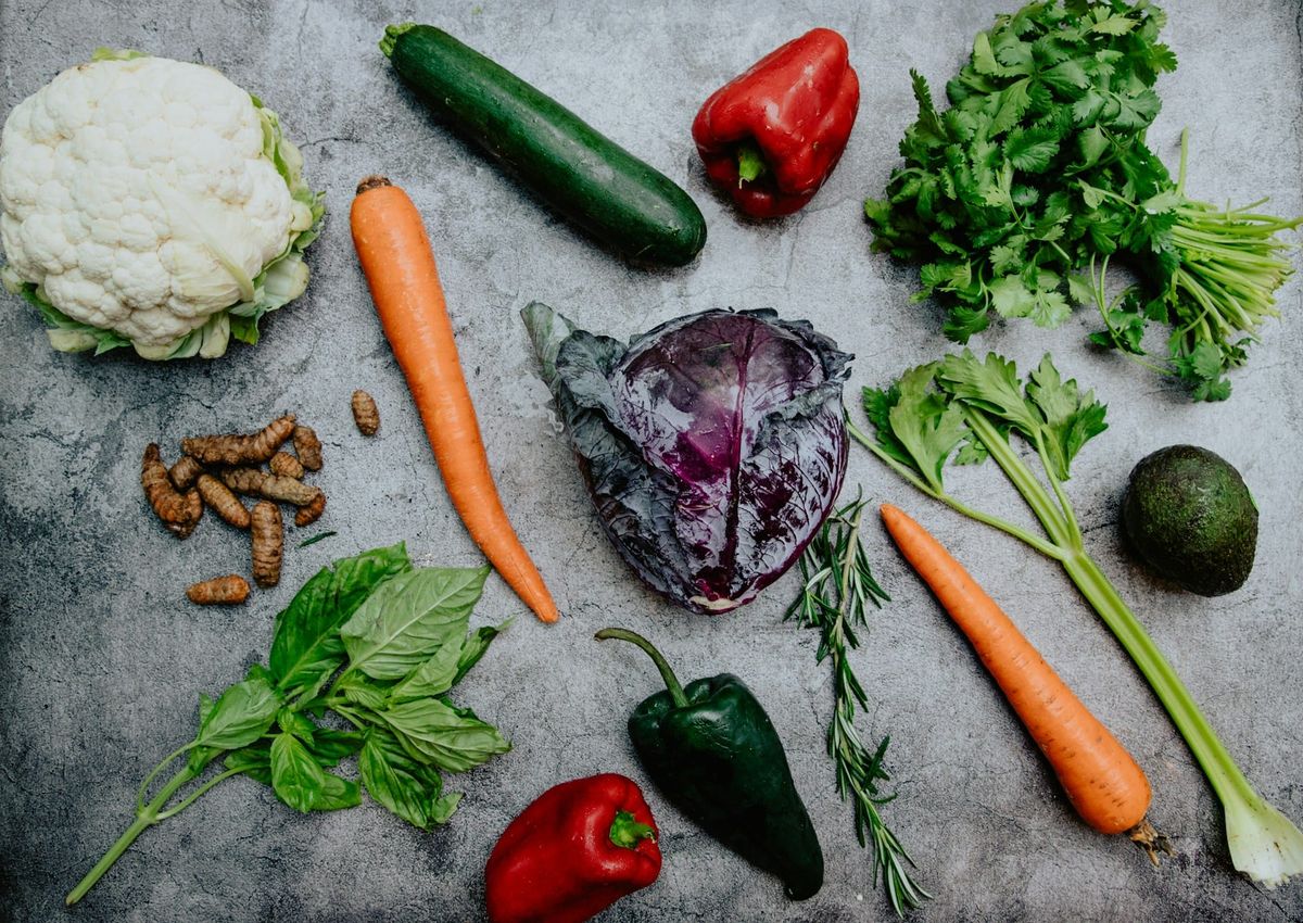 Flat lay arrangement of fresh whole vegetables and herbs including carrots, red cabbage, cauliflower, zucchini, peppers, celery, avocado, ginger, and basil on a dark stone background.