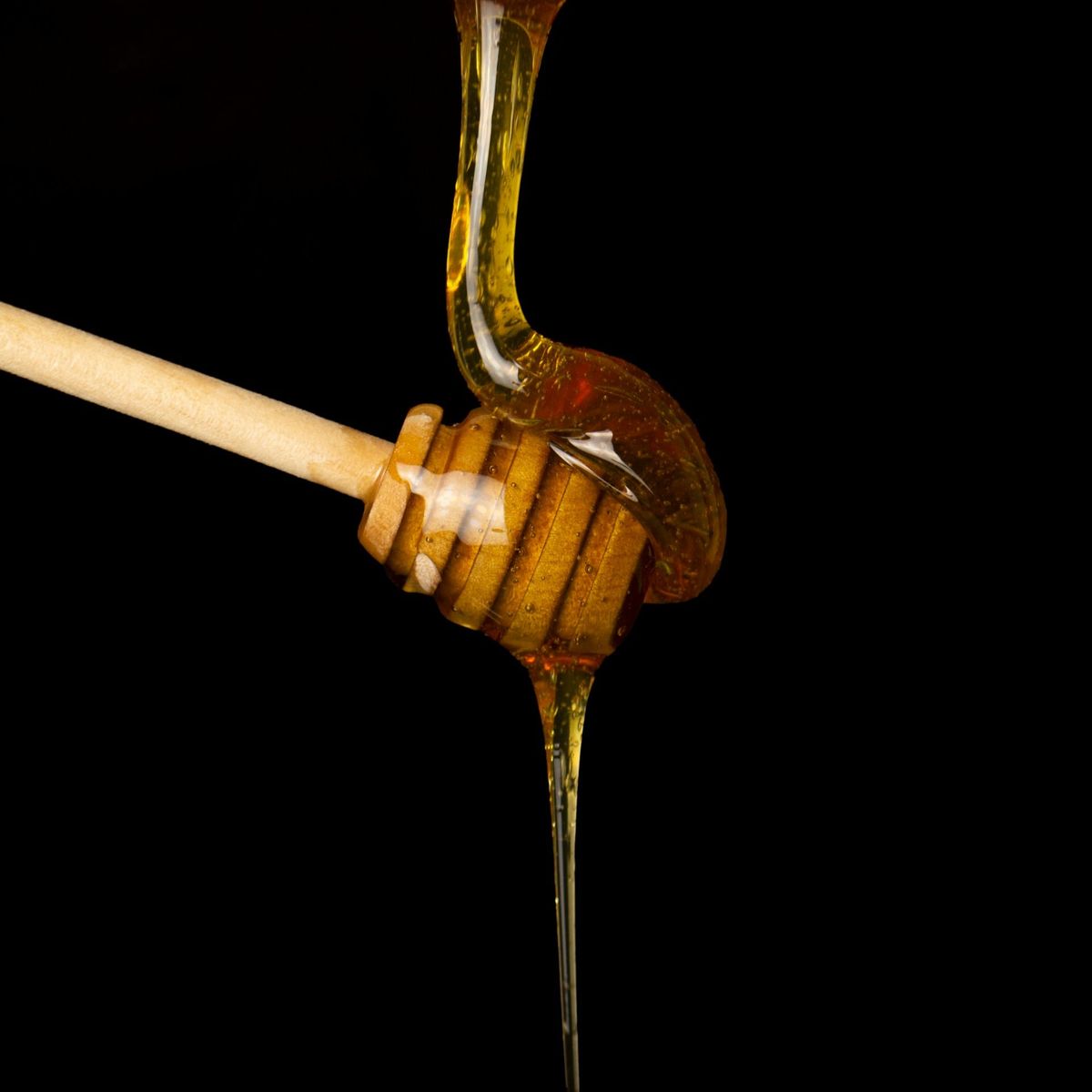 Studio macro shot on a solid black background showing thick, golden honey slowly dripping from a stream onto a ribbed wooden honey dipper.