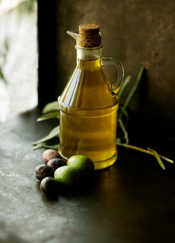 A small glass jug filled with golden olive oil sealed with a cork stopper, resting next to fresh whole olives and olive leaves on a dark surface.