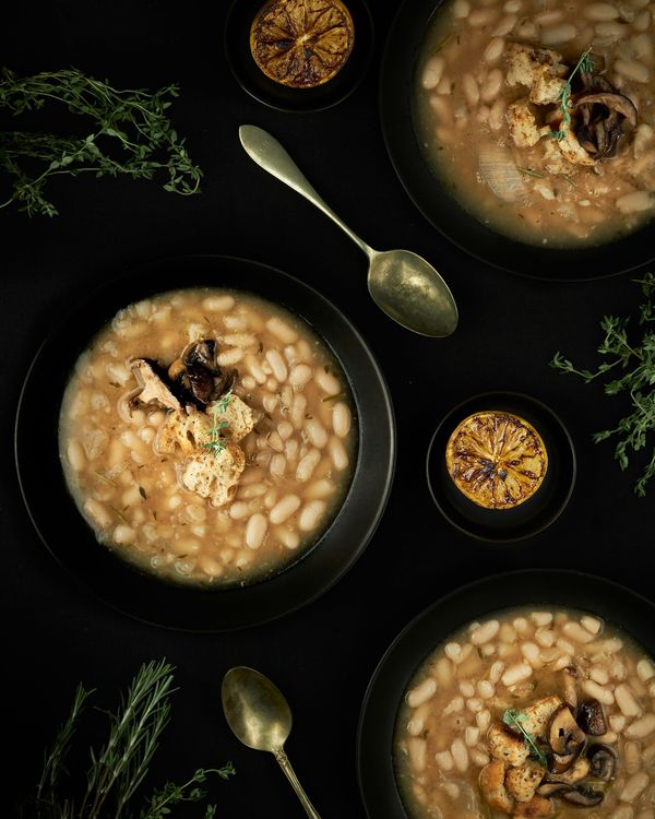 Top-down view of black bowls filled with warm white bean stew topped with mushrooms, croutons, and thyme, set against a dark background with charred lemons and brass spoons.
