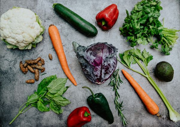 Flat lay arrangement of fresh whole vegetables and herbs including carrots, red cabbage, cauliflower, zucchini, peppers, celery, avocado, ginger, and basil on a dark stone background.