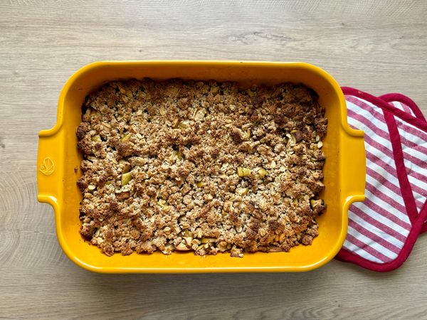 Top-down view of freshly baked apple crumble with a golden almond-oat streusel topping in a bright yellow ceramic casserole dish on a wooden surface next to a striped kitchen towel.