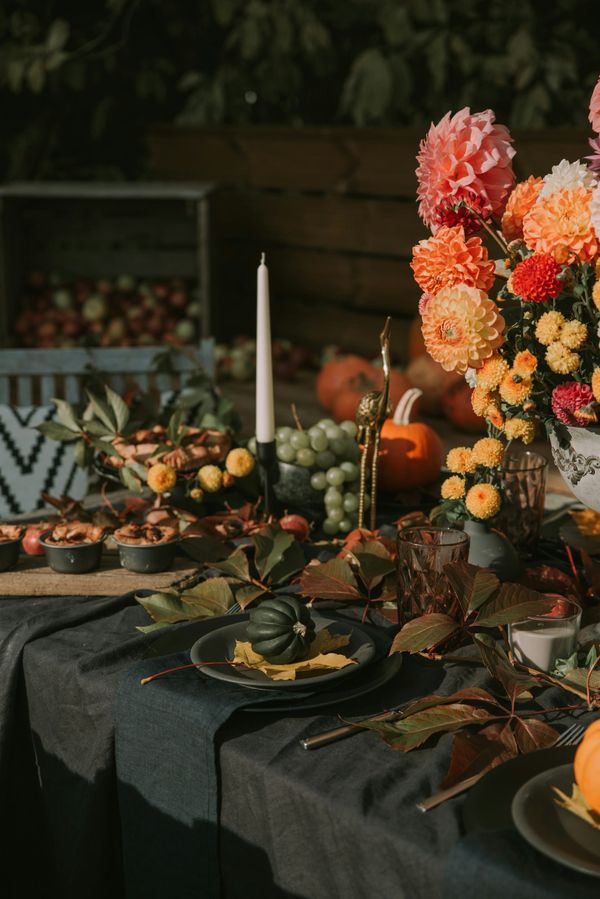 An outdoor autumn harvest table setting with dark table linens, dark grey plates, orange dahlias in a vase, taper candles, small pumpkins, and bowls of green grapes.
