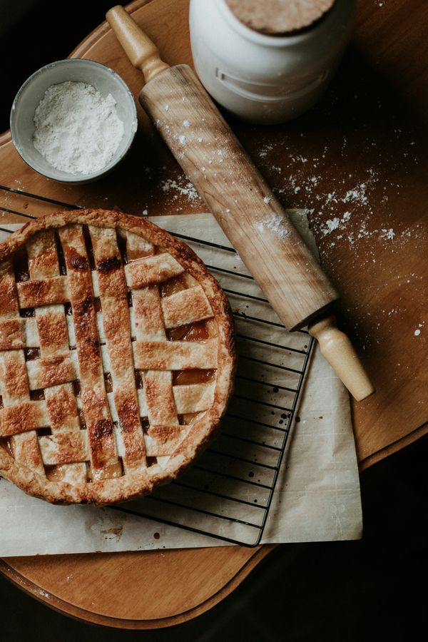 Overhead view of a freshly baked golden-brown lattice apple pie on a wire cooling rack next to a wooden rolling pin and flour bowl.