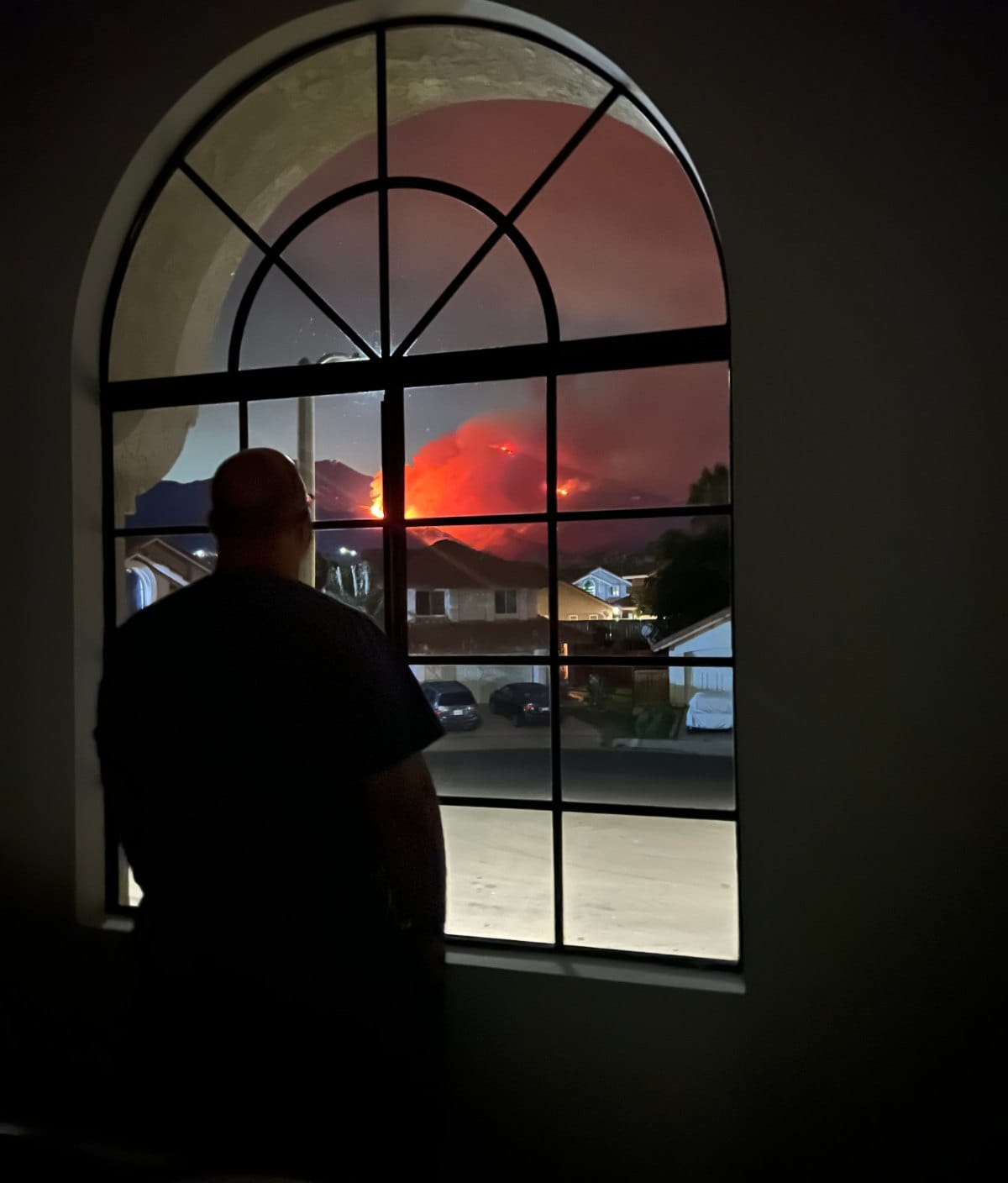 A person silhouetted at an arched window at night watches the orange glow of a nearby wildfire over rooftops.