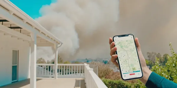 A person checks a wildfire map on their phone as smoke rises over a nearby neighborhood.