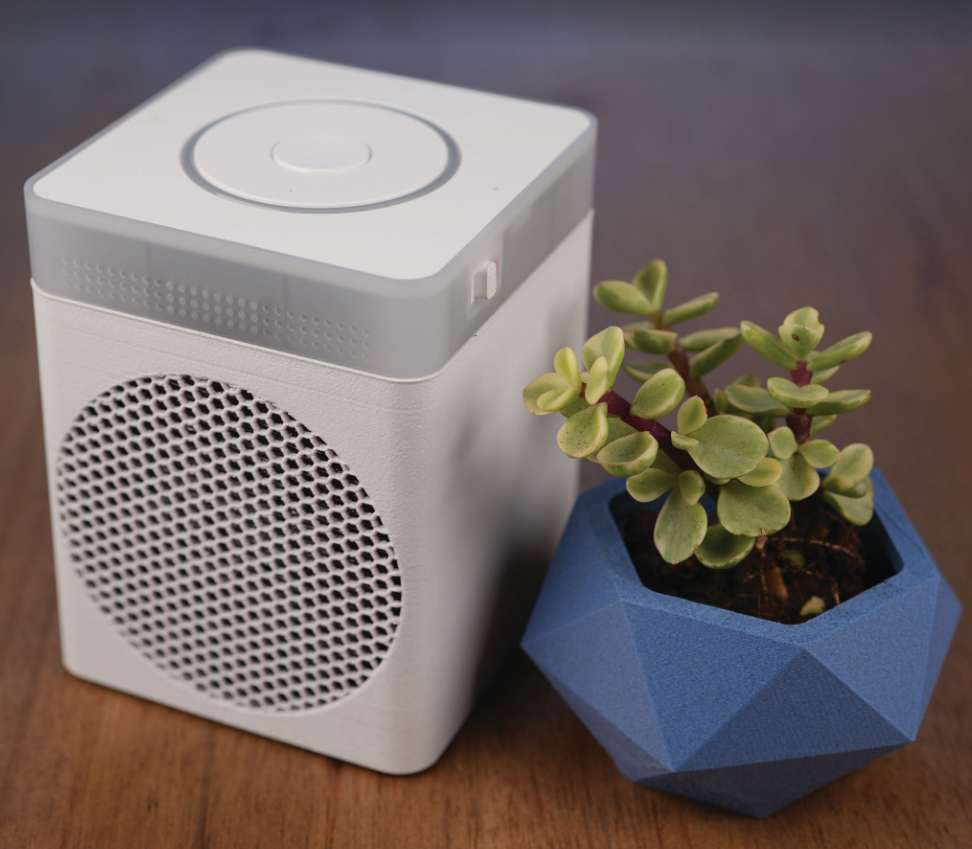A small cube-shaped speaker with a Home Assistant Voice Preview Edition on top, next to a small houseplant.