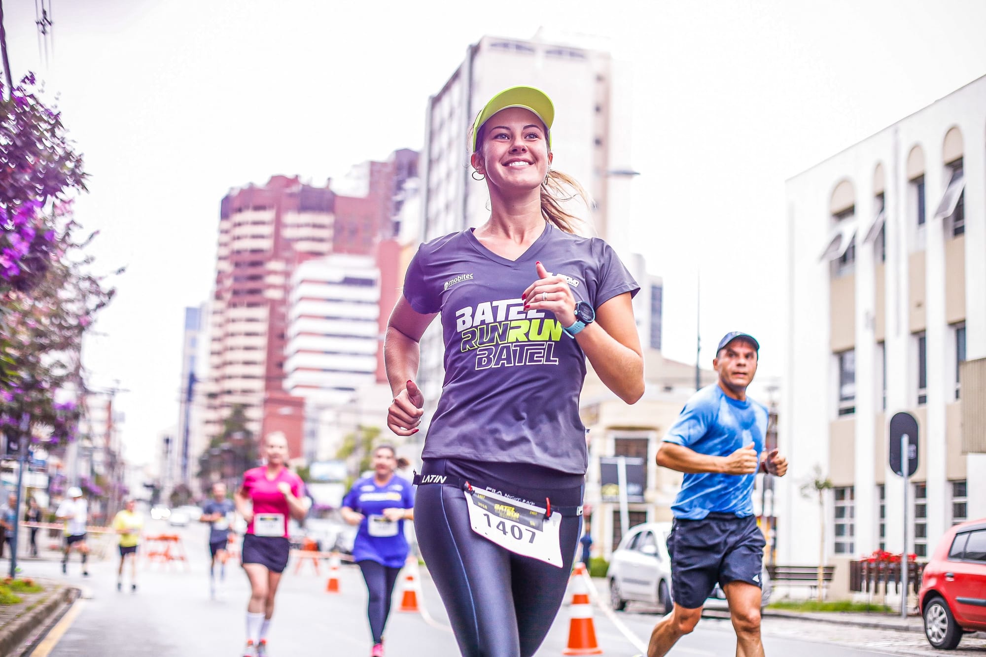 Mulher sorridente corre em avenida urbana durante a Batel Run, usando camiseta do evento e número de peito,  ao lado de outros participantes.