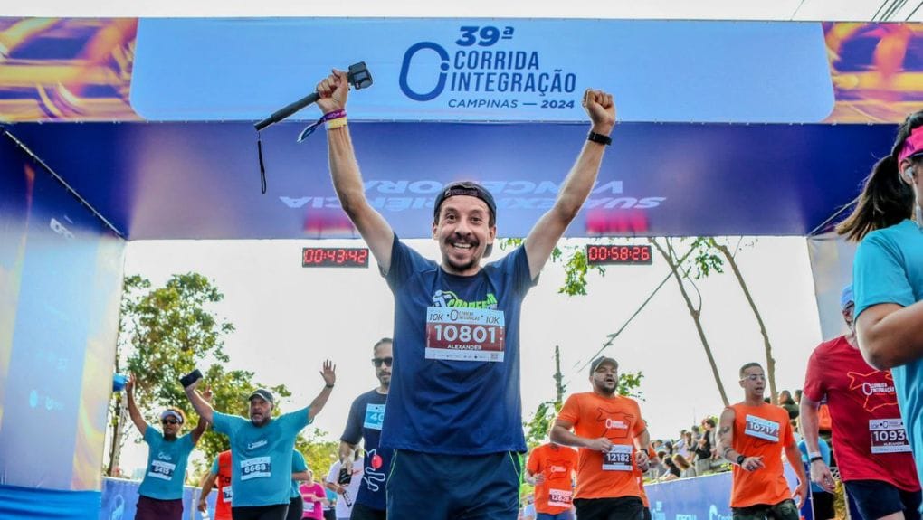 Homem comemora com os braços erguidos ao cruzar a linha de chegada da 39ª Corrida Integração em Campinas, rodeado por outros corredores.