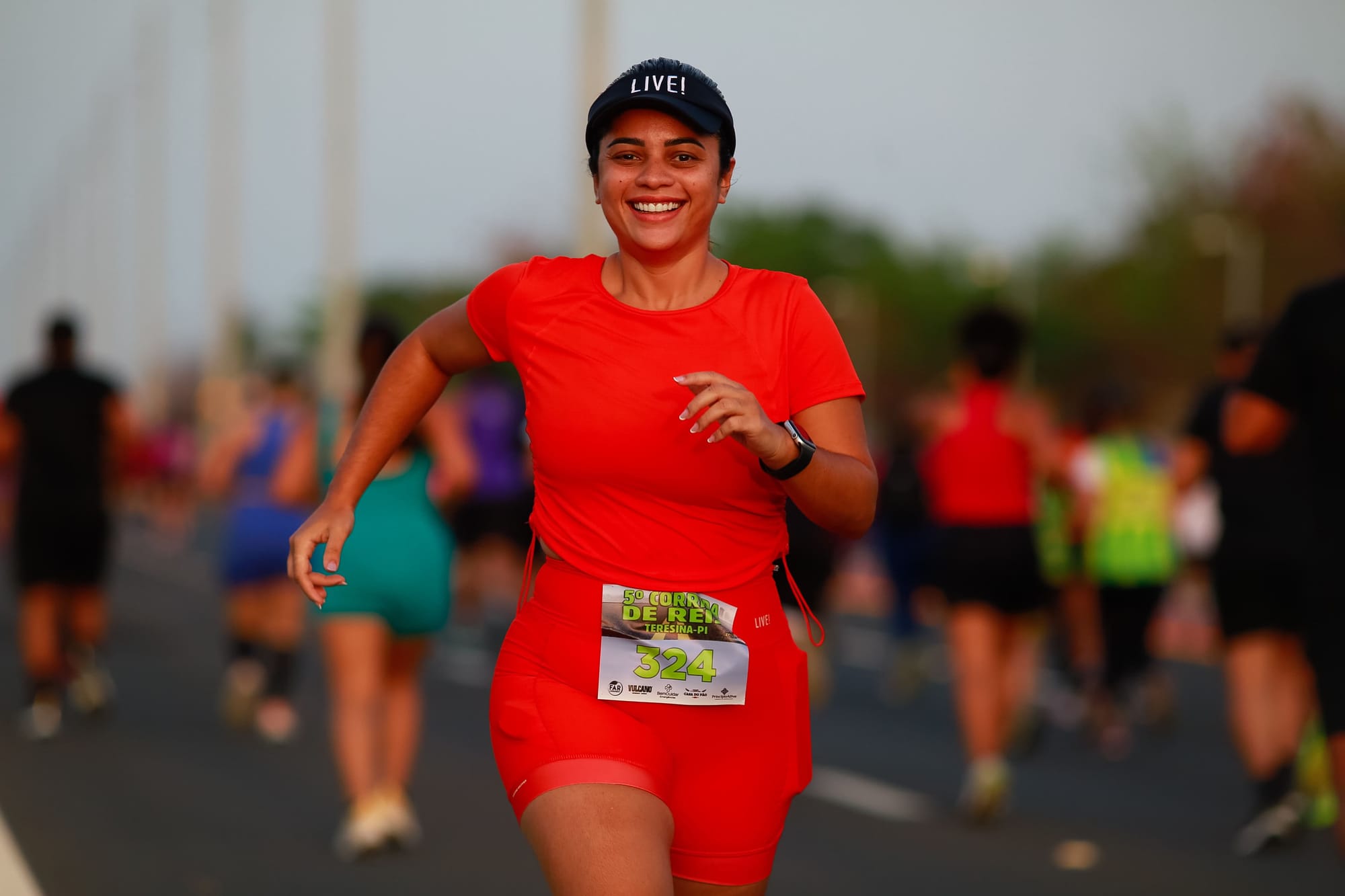 Mulher sorridente corre com roupa vermelha e número de peito durante a 5ª Corrida de Reis em Teresina, ao lado de outros participantes.