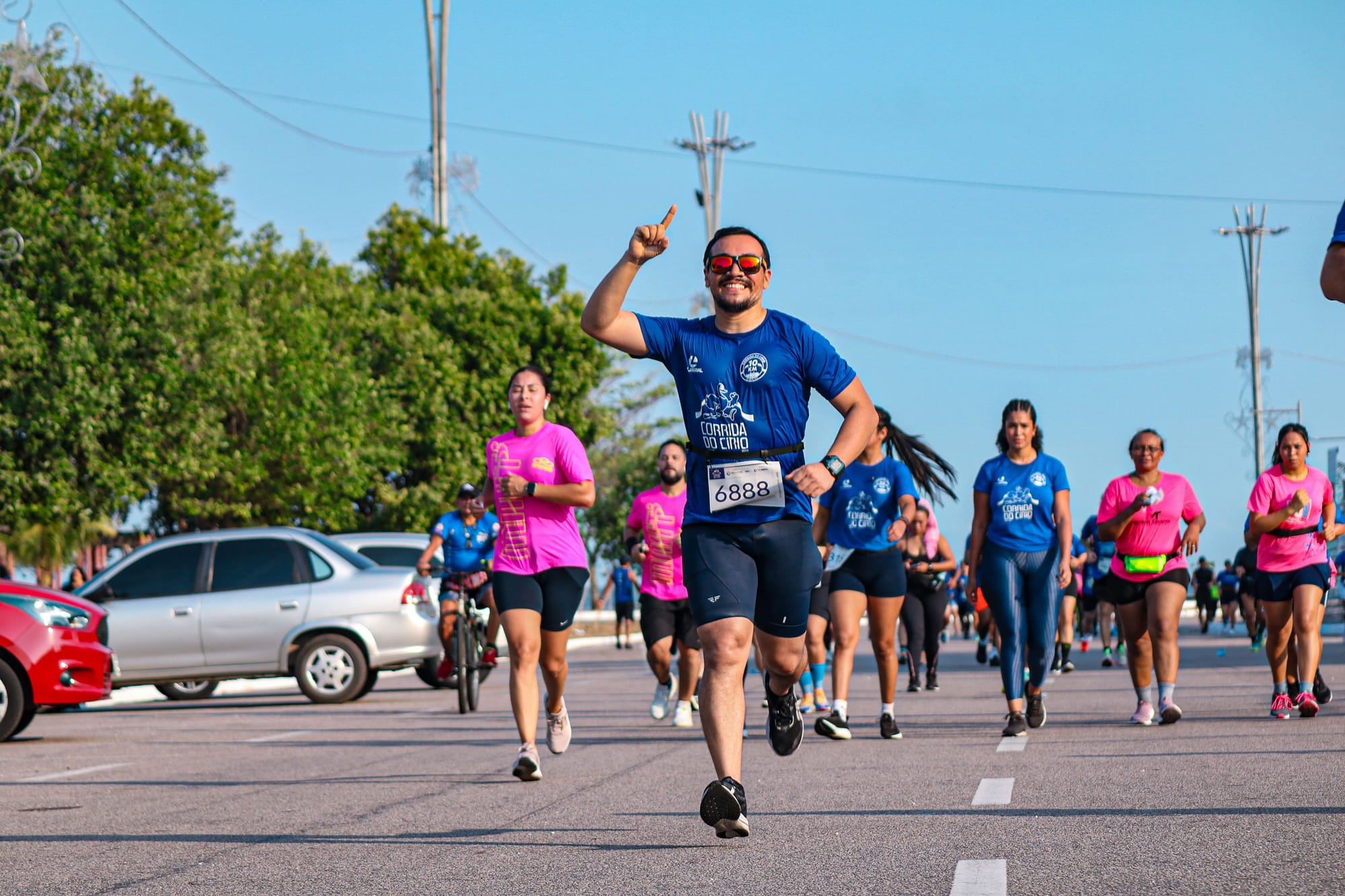 Homem sorridente corre apontando para cima durante a Corrida do Círio, rodeado por outros participantes em camiseta azul e rosa.