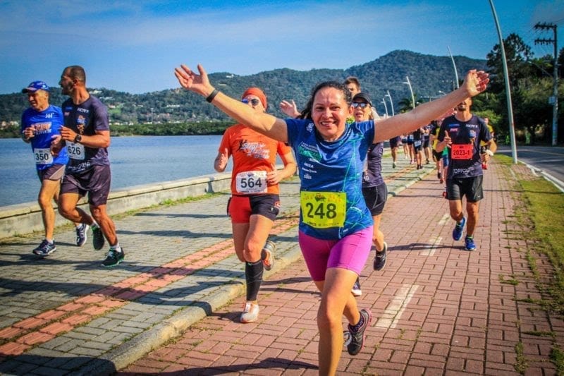 Mulher com número de peito corre sorrindo de braços abertos na Volta à Lagoa da Conceição, cercada por outros atletas e paisagem com montanhas ao fundo.