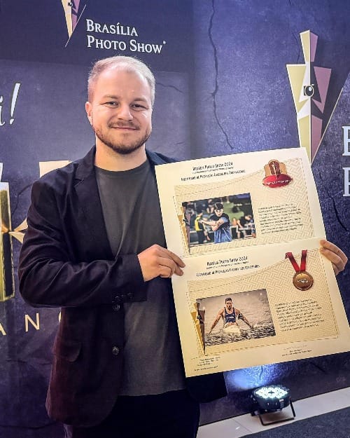 Fotógrafo Gabriel Haesbaert sorri ao segurar certificado de premiação do Brasília Photo Show 2024 com fotos premiadas em destaque.