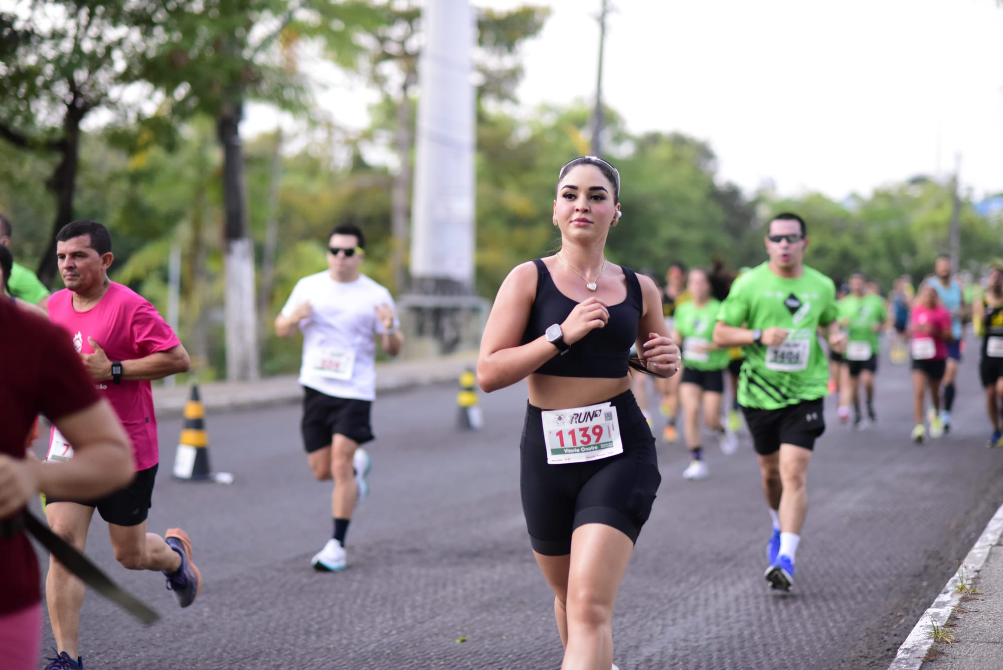 Mulher com número de peito corre em prova de rua ao lado de outros atletas, durante o Pátio Gourmet Run, em avenida arborizada.