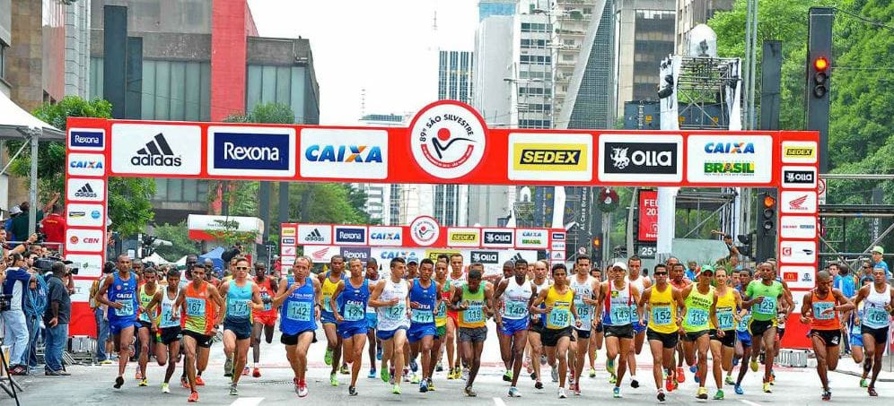 Corredores largam na Avenida Paulista durante a São Silvestre, passando sob o pórtico com marcas de patrocinadores.