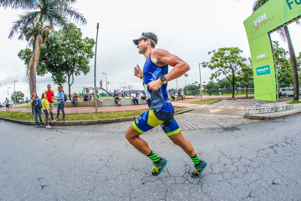 Homem corre com roupa de triatleta durante prova da Volta Internacional da Pampulha, com parque e espectadores ao fundo.