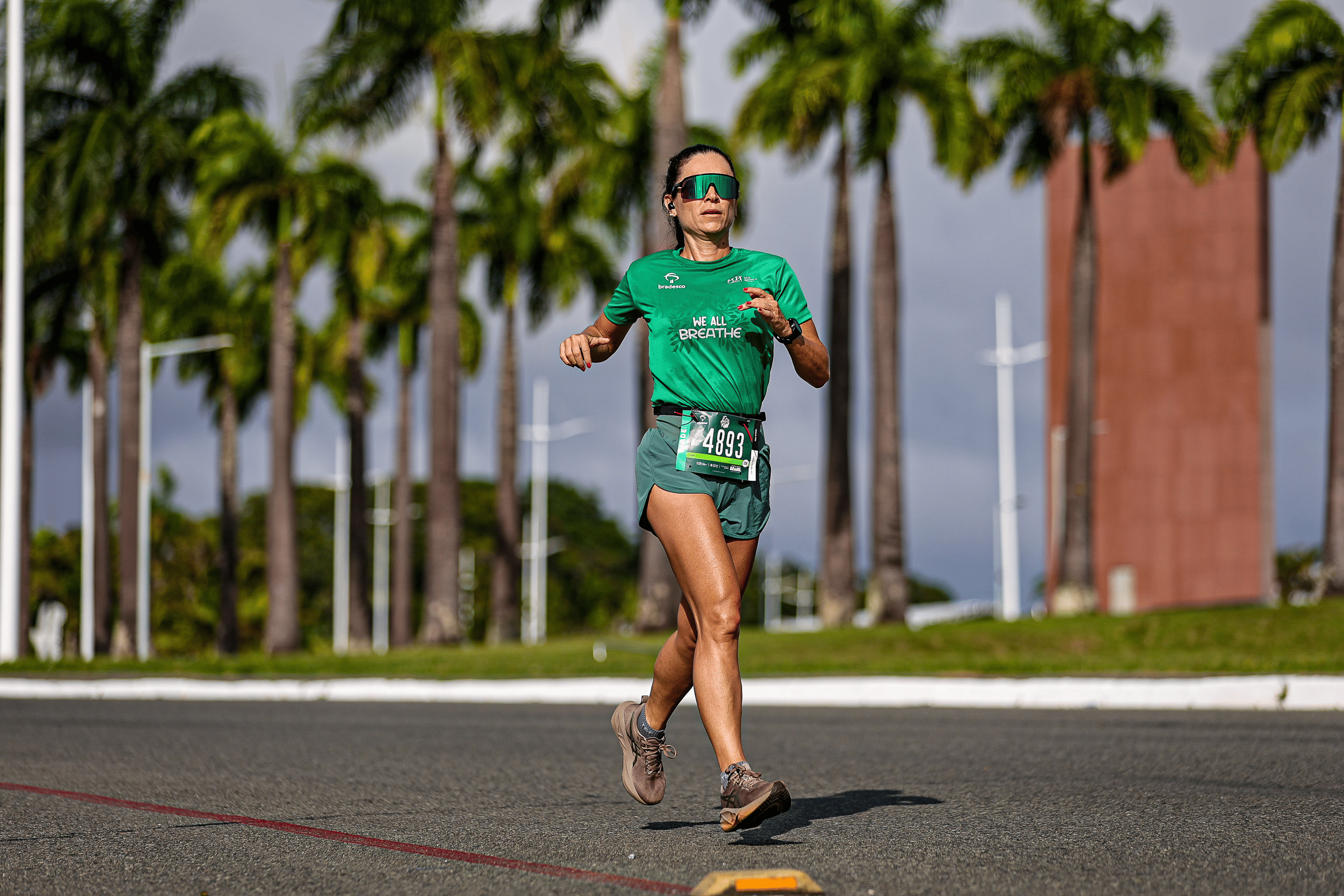Mulher corre sozinha em uma avenida durante uma prova de rua. Ela veste camiseta e shorts verdes, usa óculos esportivos espelhados e fones de ouvido.