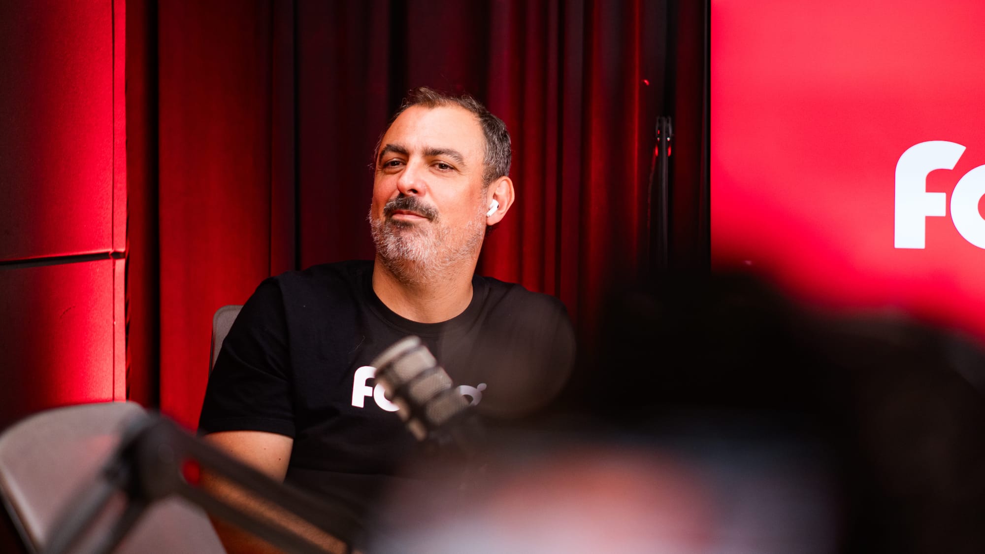 Homem de barba e camiseta preta com a logo da Fotto participa de uma live. Ele está sentado em frente a um microfone, usando um fone de ouvido branco, com expressão confiante