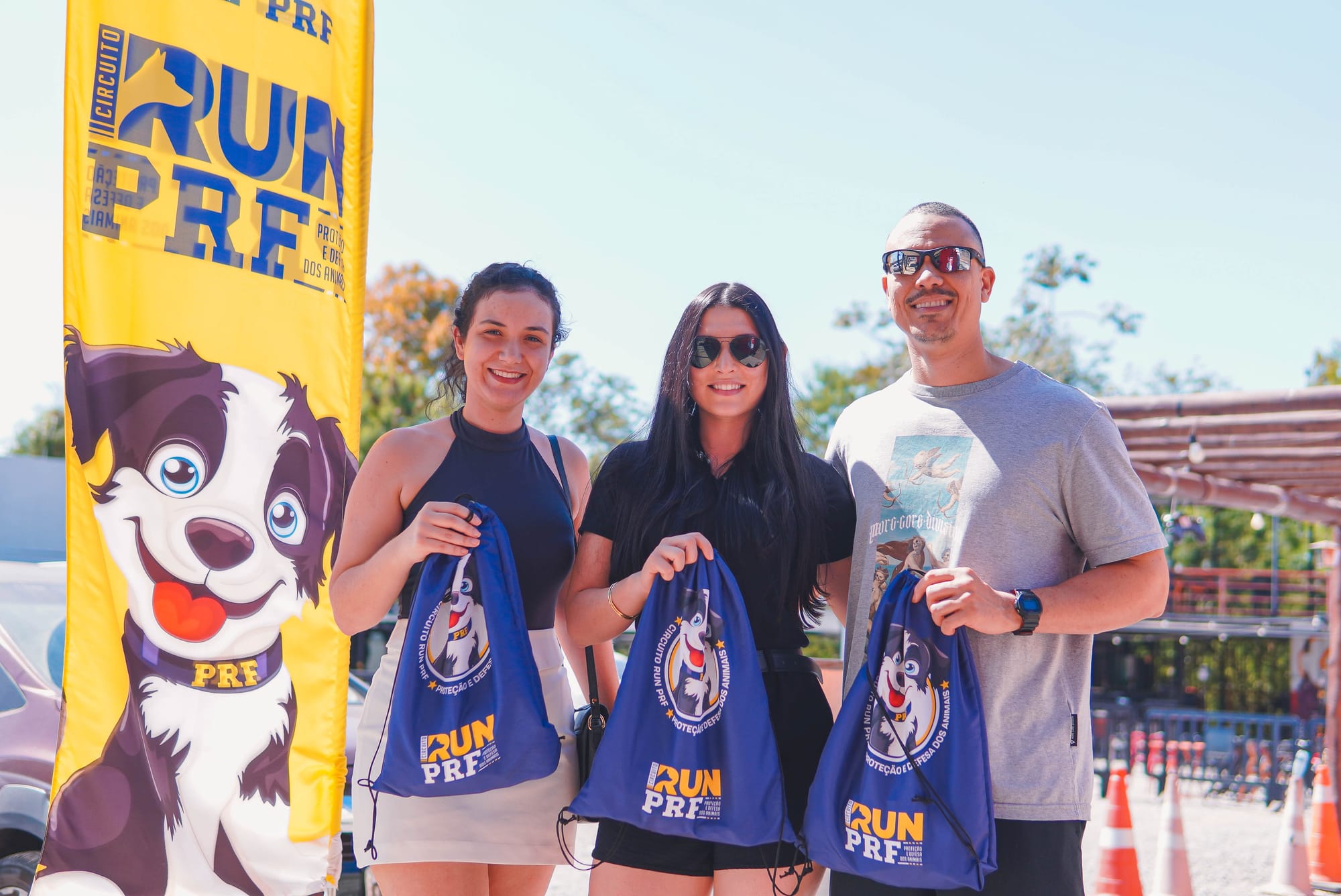 rês pessoas posam sorridentes para a foto ao ar livre, segurando o kit para a corrida Circuito Run PRF